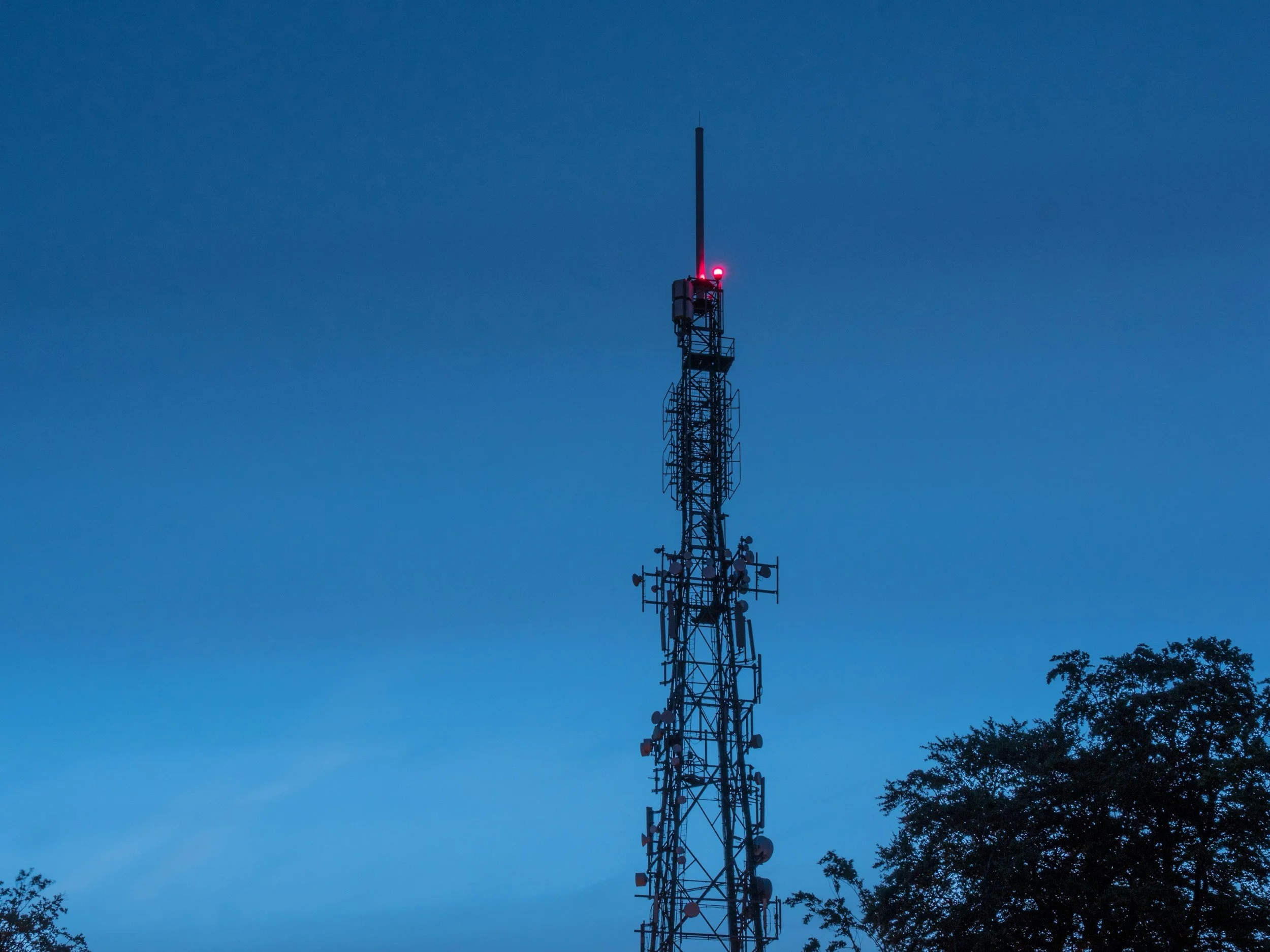 A tall radio tower with antennas and equipment, illuminated by a red light at the top, against a dusky sky with silhouettes of trees.