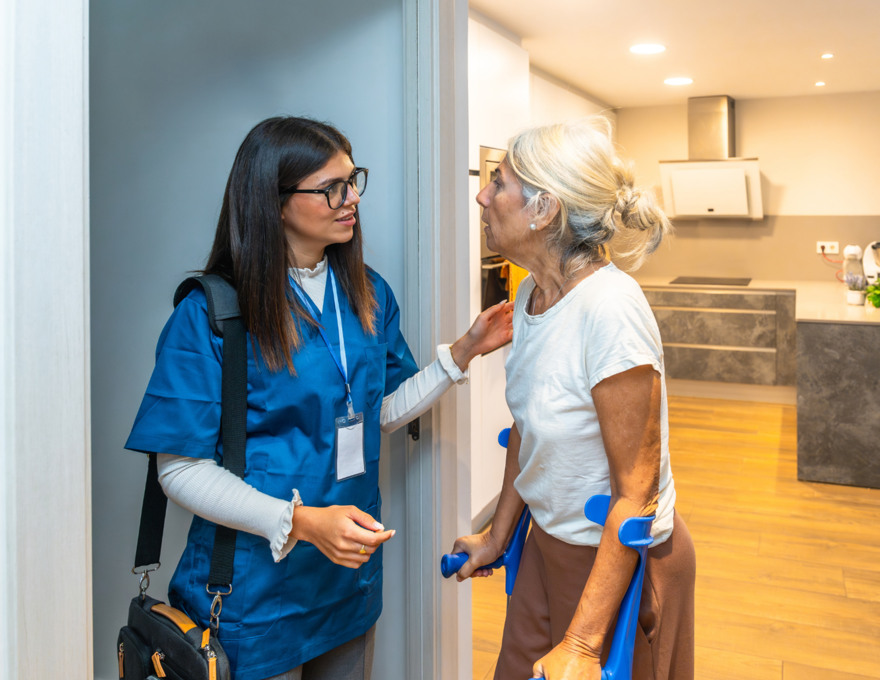 A young female nurse in scrubs and glasses comfort a senior woman using crutches, in a residential setting.