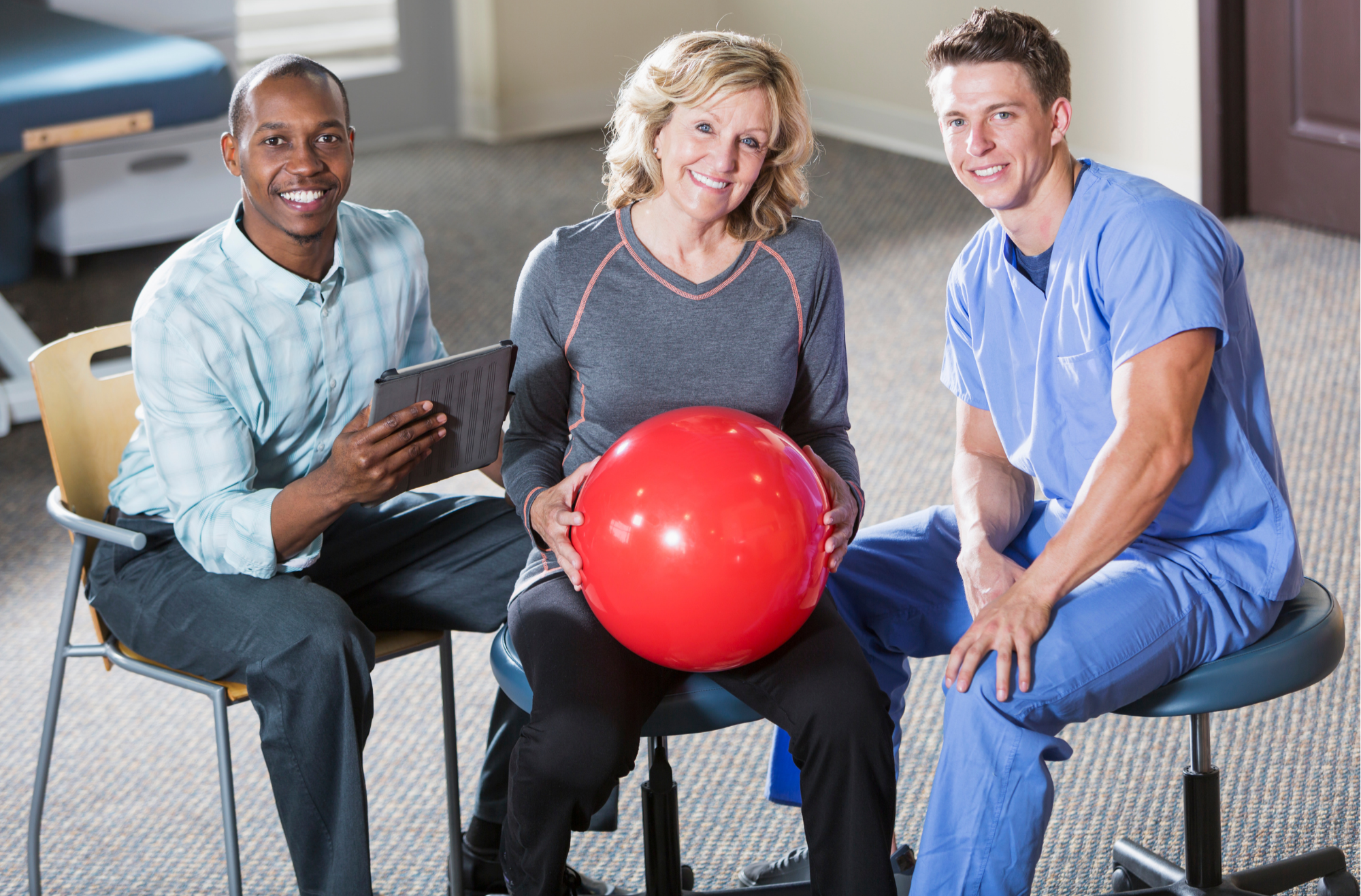 A middle-aged woman holding a red exercise ball, seated between a man in business attire with a tablet and a young male healthcare worker in blue scrubs, in a therapy or rehabilitation setting.