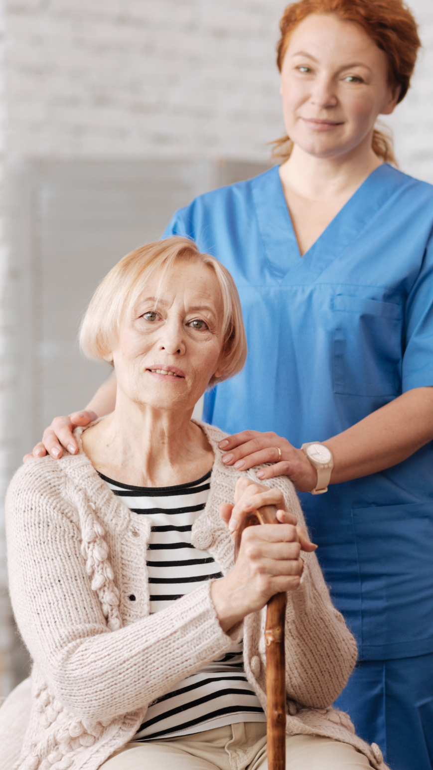 A nurse or caregiver in blue scrubs standing beside an elderly woman with short blonde hair, wearing a beige cardigan and black-and-white striped shirt, holding a walking cane, in a healthcare setting.