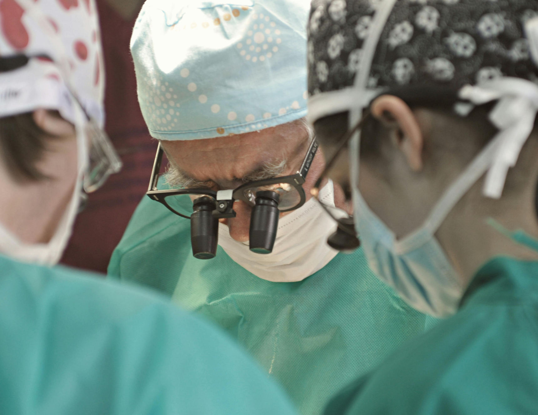 Three surgeons in operating room performing surgery, wearing scrubs, masks, and head coverings.