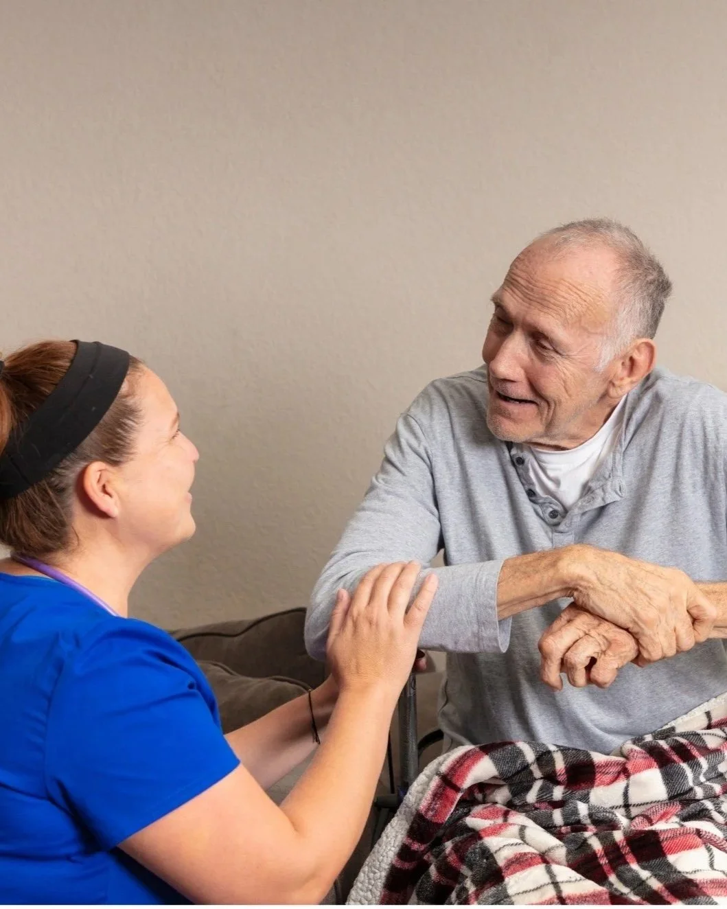 A female healthcare worker smiling and holding the hand of an elderly man sitting in a wheelchair, wrapped in a plaid blanket, in a room with a beige wall.
