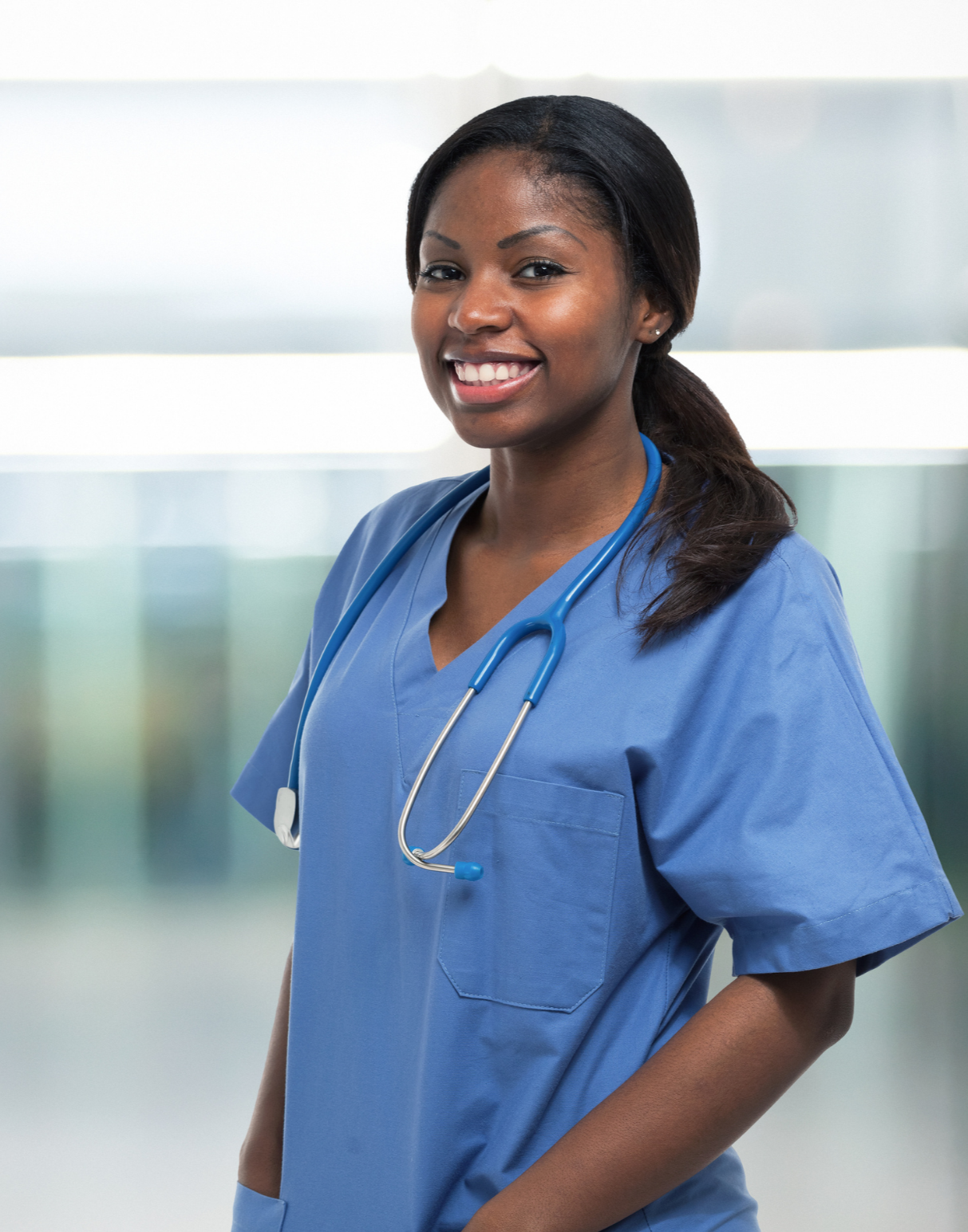 Young woman nurse smiling wearing blue scrubs and stethoscope on her neck in a healthcare setting.