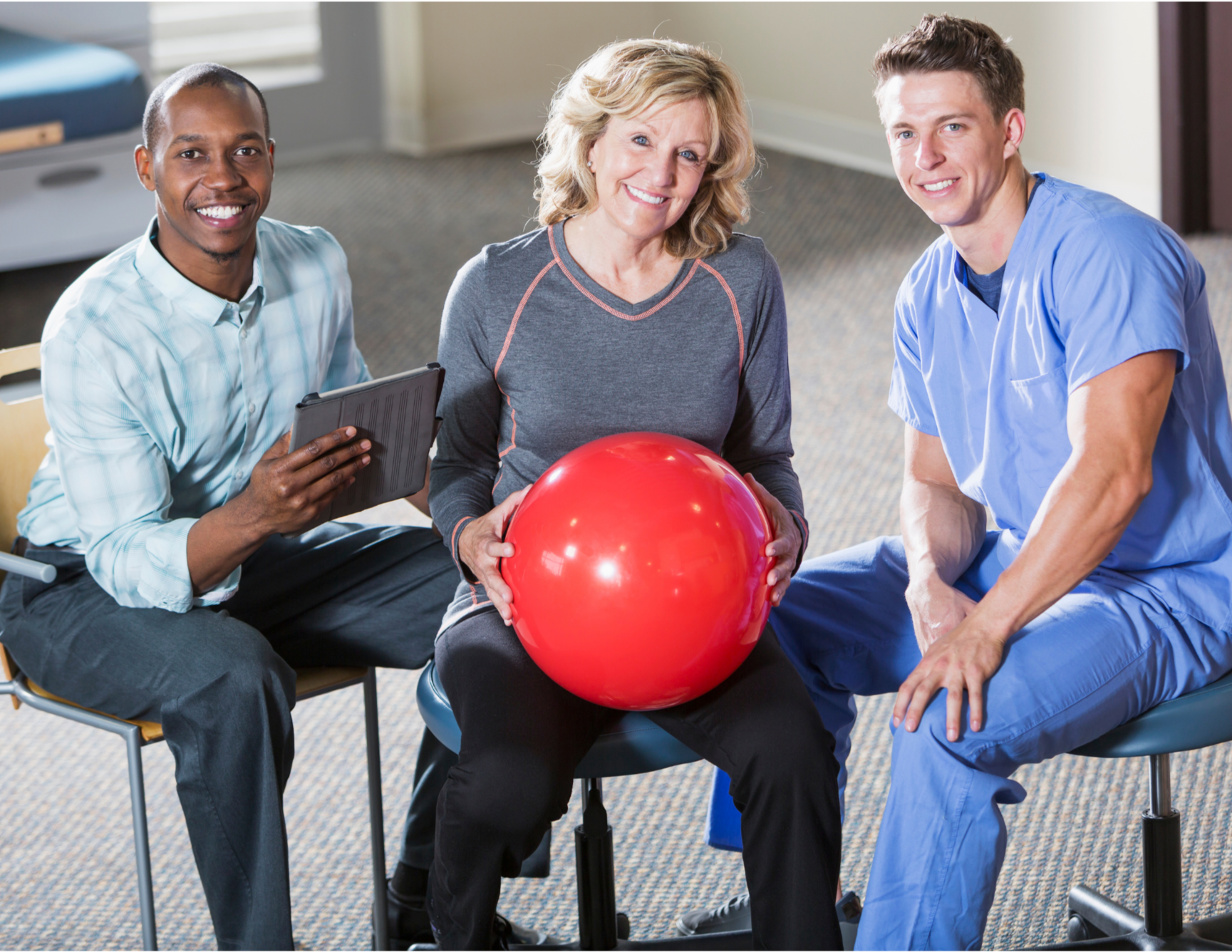 A woman participating in physical therapy with a red exercise ball, flanked by two healthcare professionals in a clinical setting.
