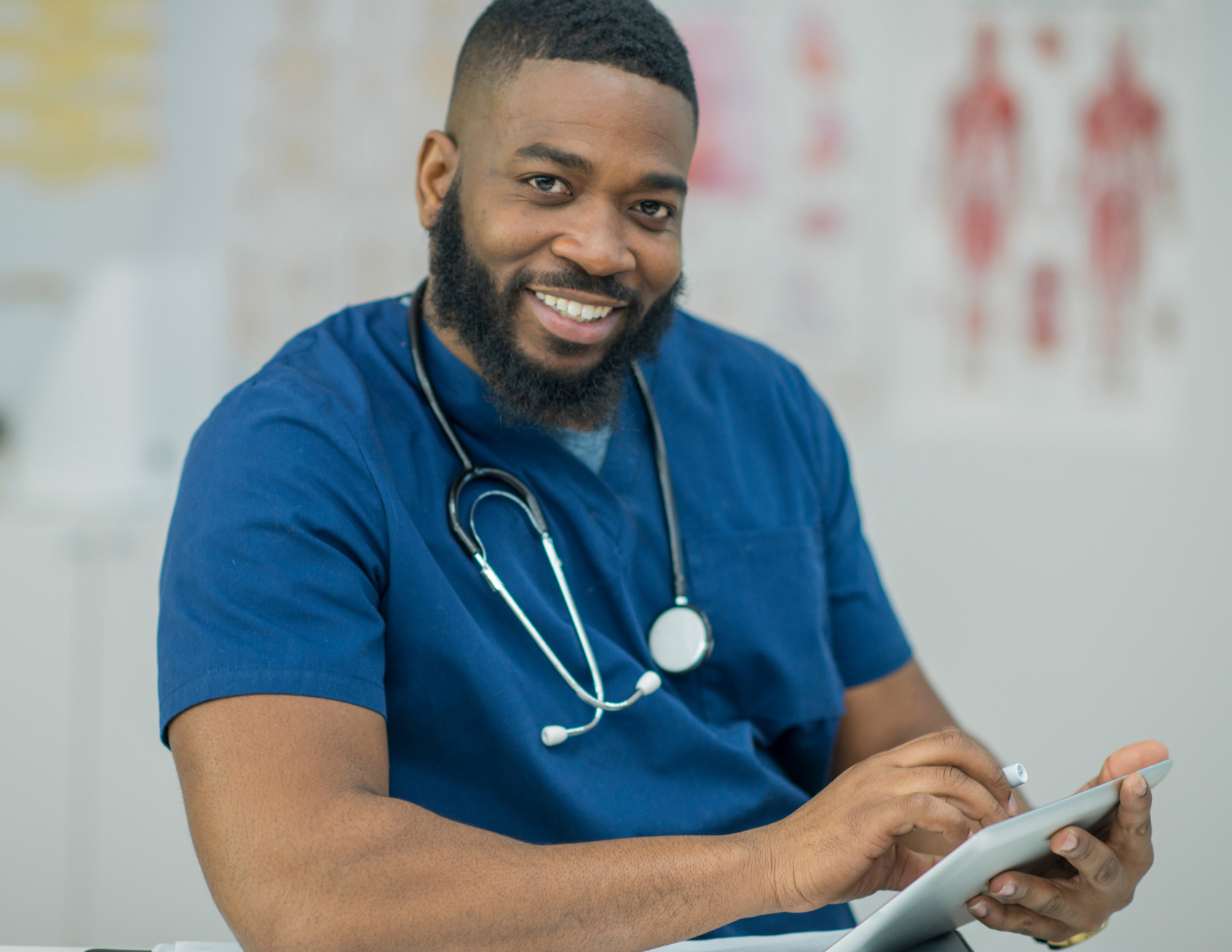 Smiling male healthcare professional with a stethoscope around his neck using a tablet in a medical setting.