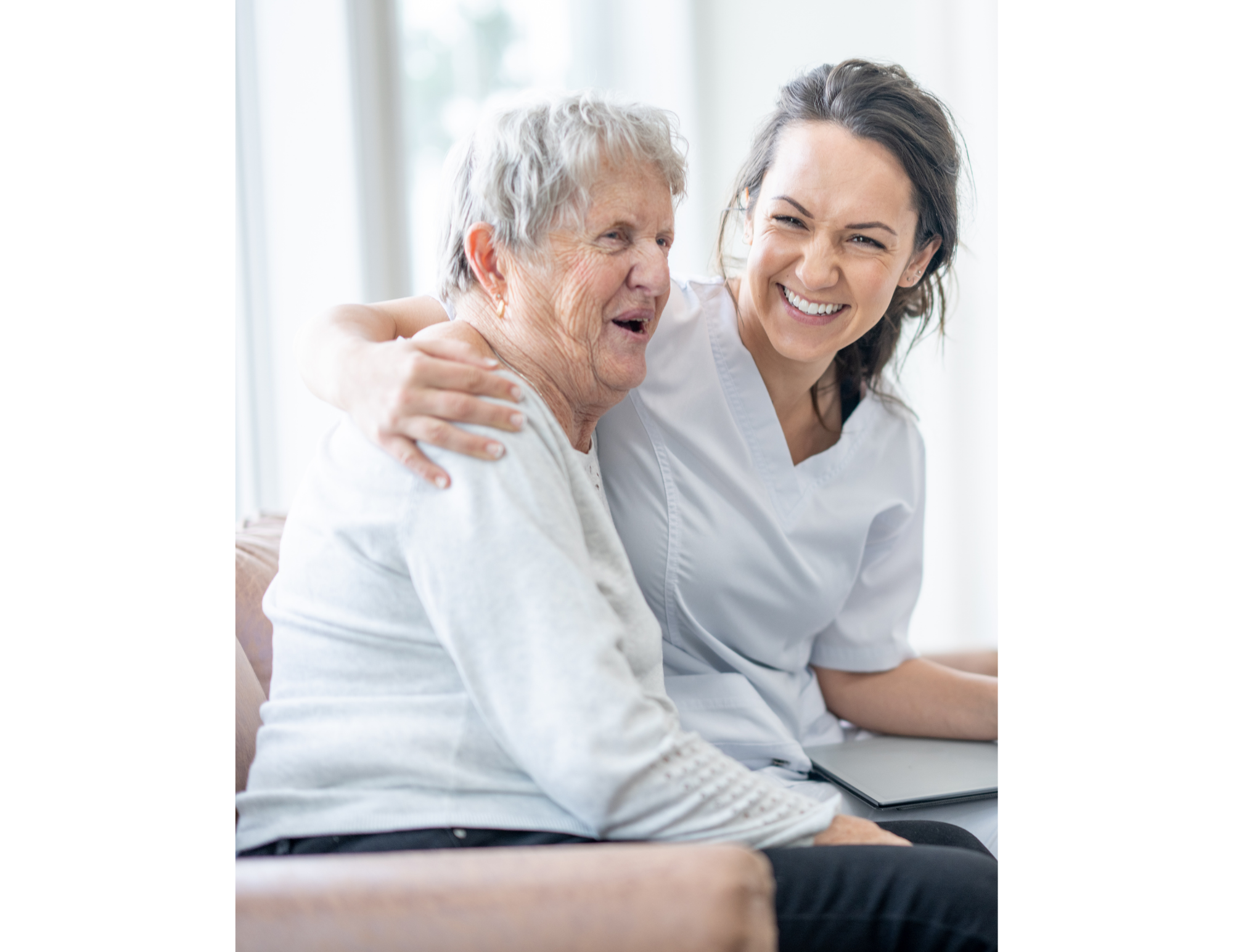 A young female caregiver with dark hair and a white uniform happily hugs an elderly woman with gray hair, both smiling, indoors with a large window in the background.
