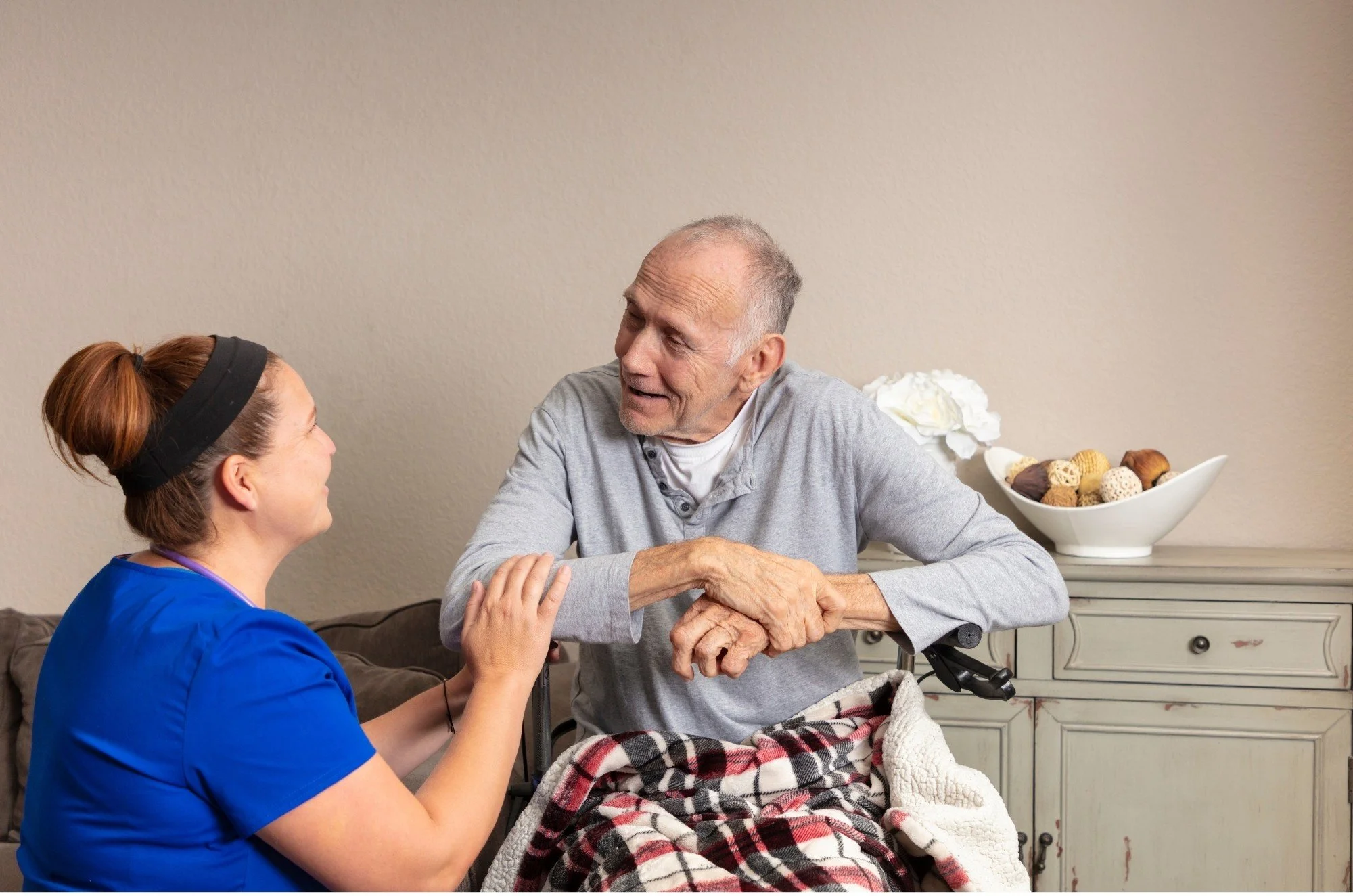 A nurse or caregiver talking and holding hands with an elderly man in a wheelchair, with a bowl of decorative balls and flowers on a table in the background.