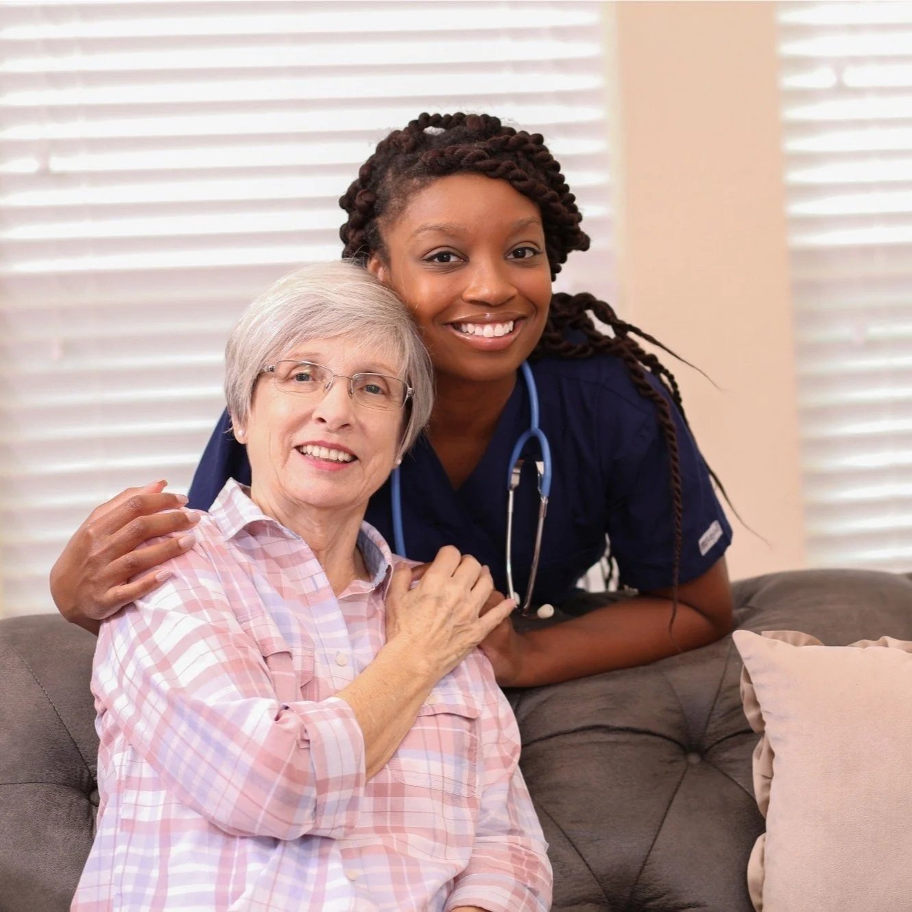 A young female nurse with a stethoscope around her neck, smiling, with an elderly woman with glasses, sitting on a couch, in a room with blinds in the background.