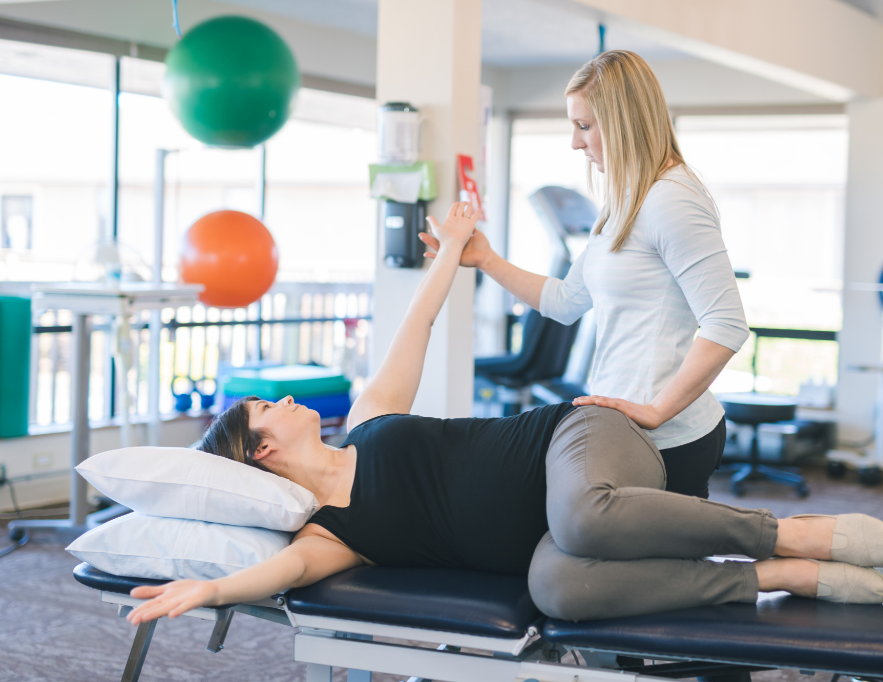 A physical therapist or chiropractor assisting a woman with a stretch or exercise on a treatment table in a rehab or therapy center with exercise balls and gym equipment in the background.
