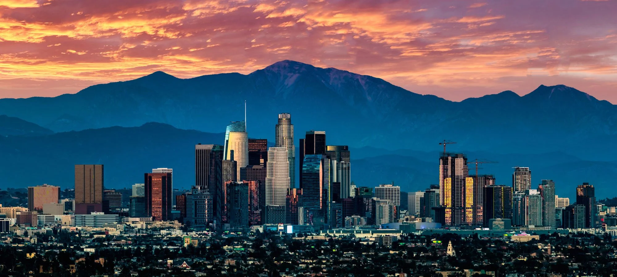Skyline of a city with skyscrapers in front of a mountain range during sunset.
