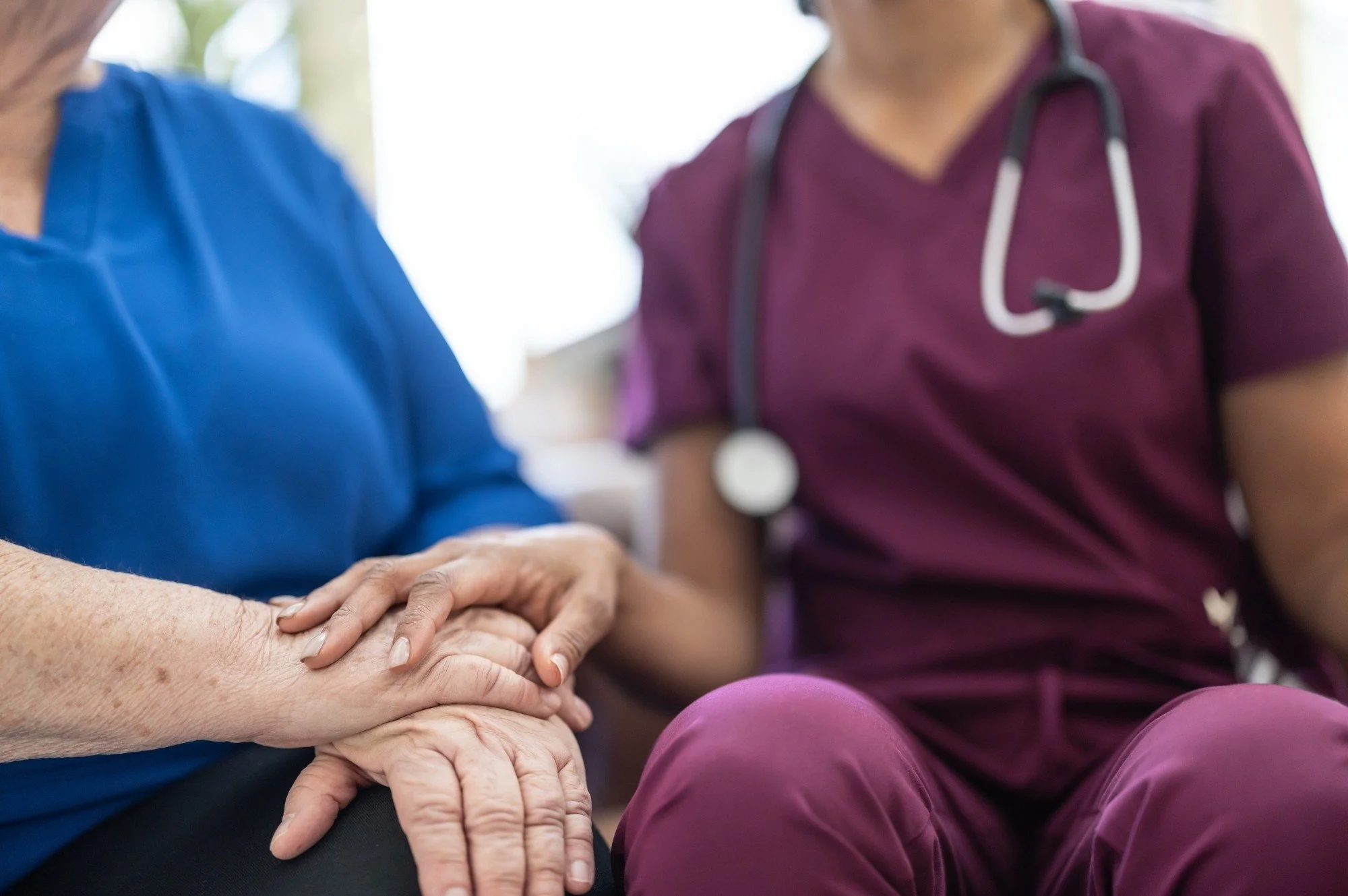A healthcare worker in purple scrubs with a stethoscope around their neck holding the hand of an elderly woman in a blue top, indicating a comforting moment.