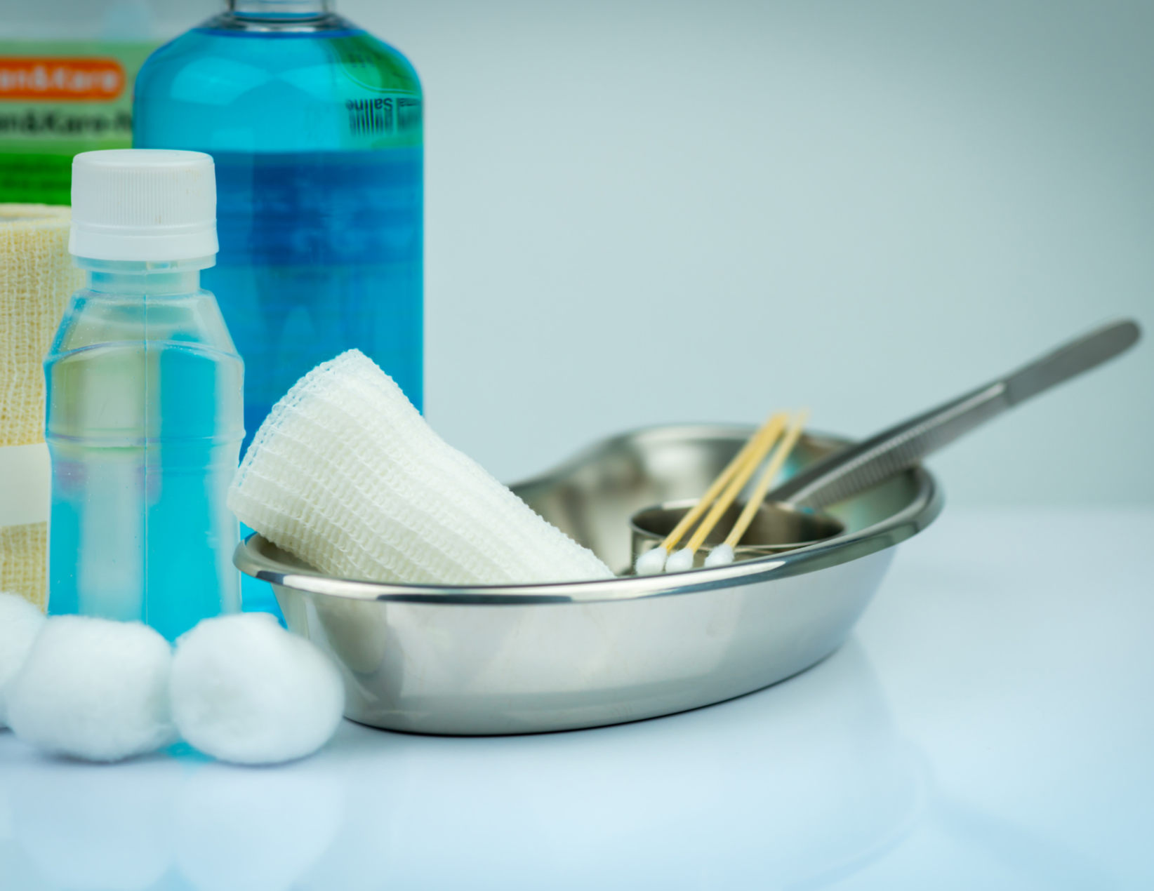 Medical supplies including a small clear bottle of antiseptic, a blue bottle, a yellow sponge, cotton balls, a metal tray with a rolled gauze, chopsticks, and a scalpel.