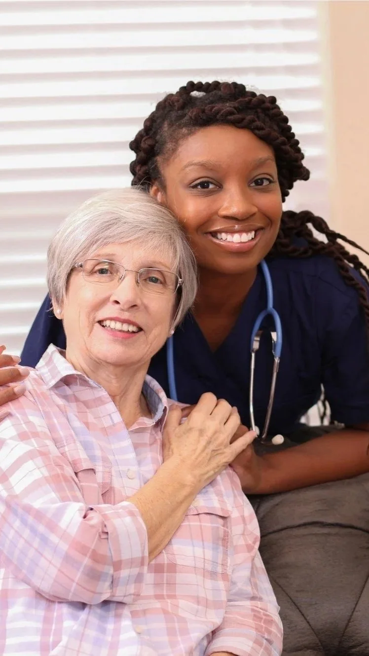 A young African American female nurse in scrubs with a stethoscope around her neck smiling and posing with an older Caucasian woman with glasses, who is also smiling and wearing a pink and white checkered shirt.