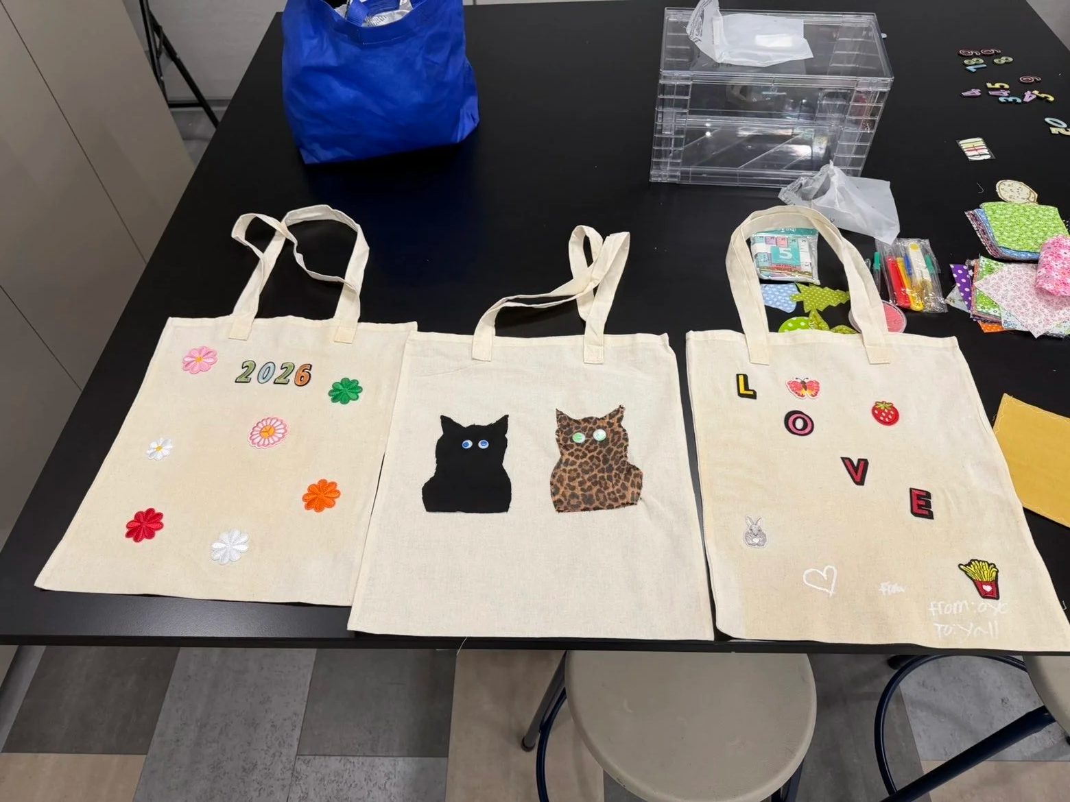 Three completed upcycled tote bags with embroidered flowers, appliquéd cats, and a "LOVE" design displayed after a workshop