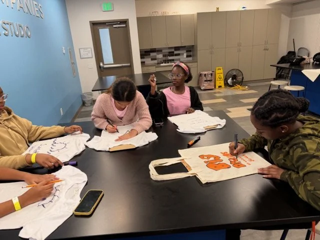 Four girls seated around a table drawing and painting designs on tote bags and t-shirts during a workshop