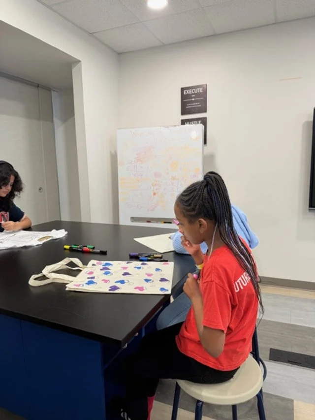 Young girl seated at a table drawing and decorating a tote bag with colorful markers during a workshop
