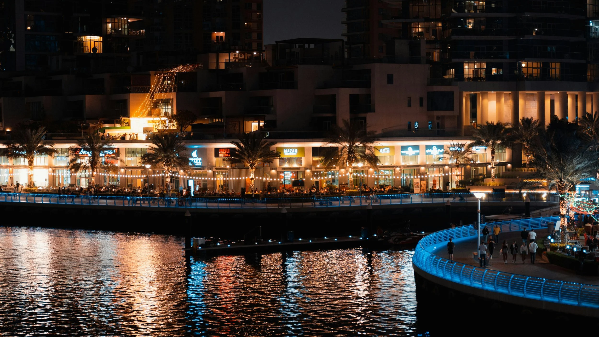 Night view of Dubai Marina with waterfront restaurants, palm trees, and people walking along the illuminated promenade reflected in the water.