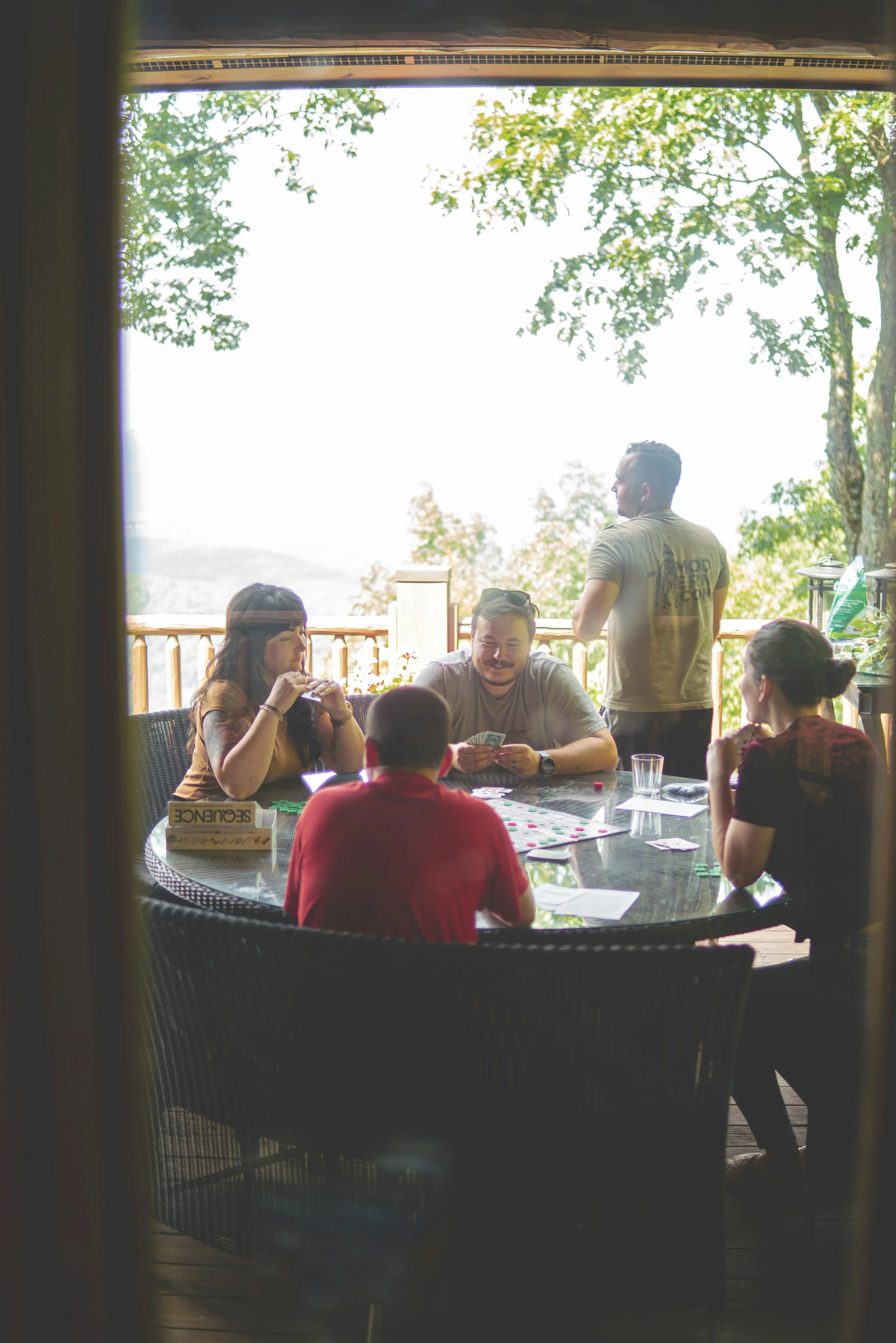 Group of friends playing a board game around a table on a covered outdoor deck at a mountain villa, surrounded by trees and scenic views.
