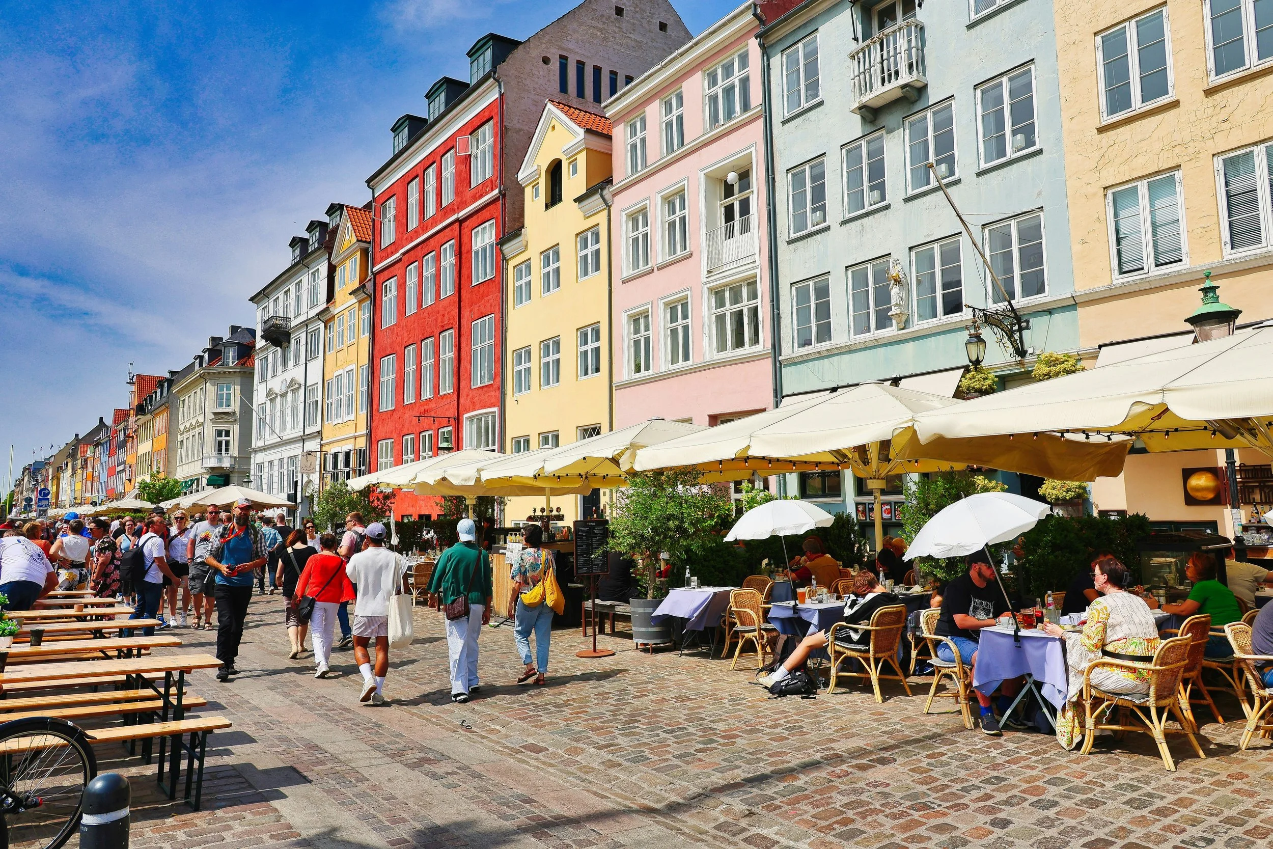 Crowded day scene in Nyhavn, Copenhagen, with colorful waterfront buildings, outdoor cafés, and tourists walking along the cobblestone street.