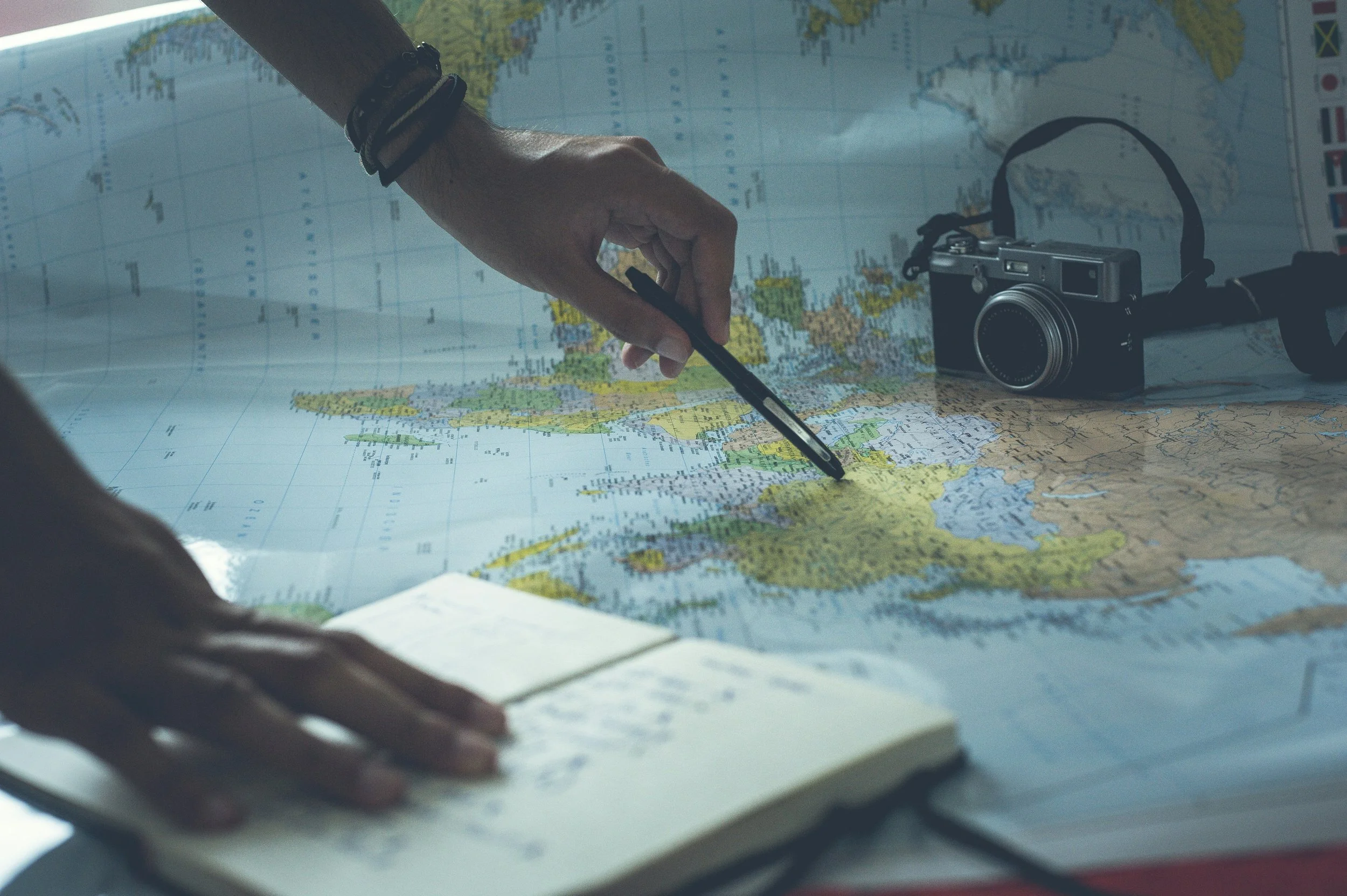Person pointing at a location on a world map with a pen while planning a trip, with a notebook and camera nearby.