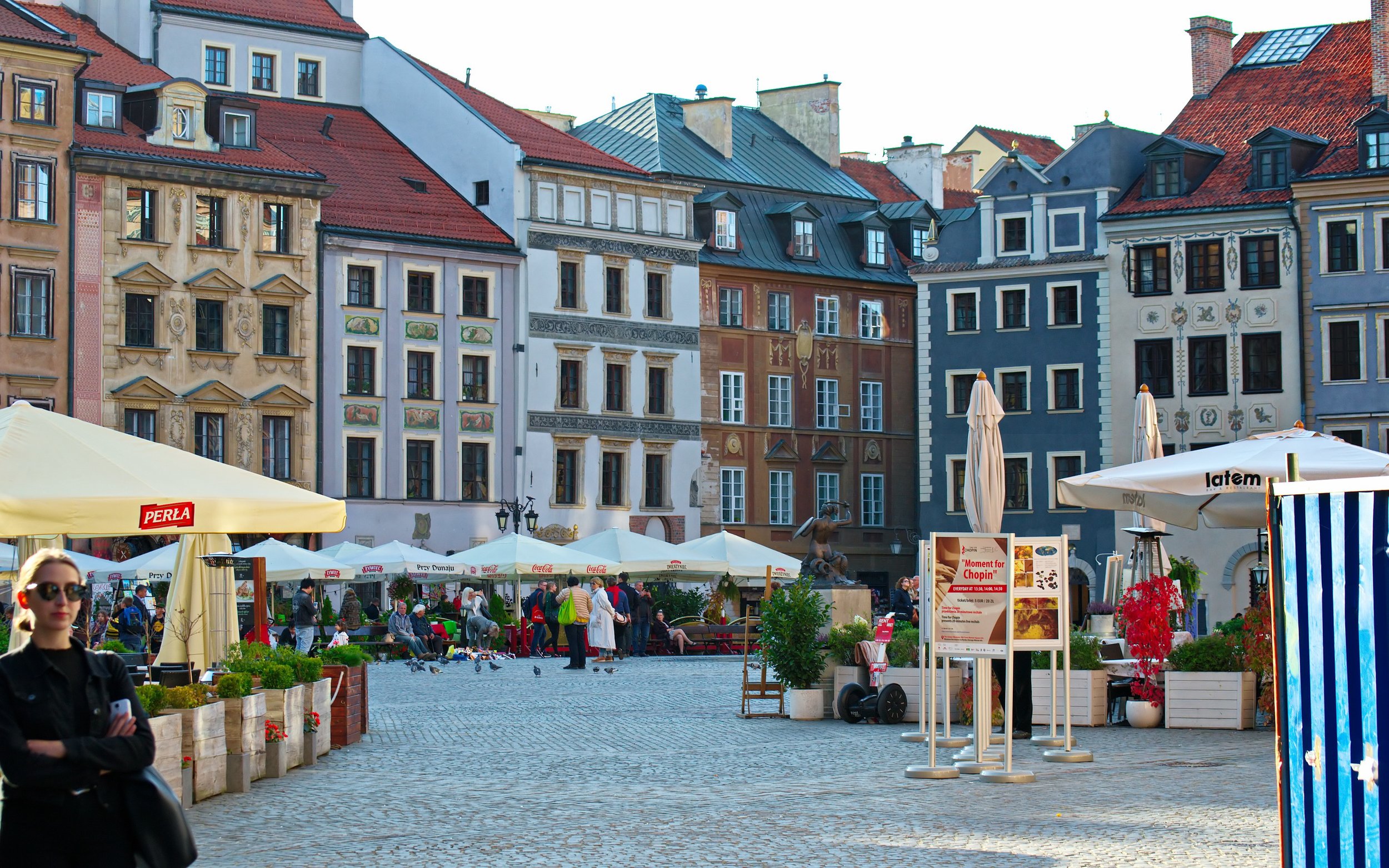 Warsaw Old Town Square with colorful historic buildings, outdoor café umbrellas, and tourists walking across the cobblestone plaza.