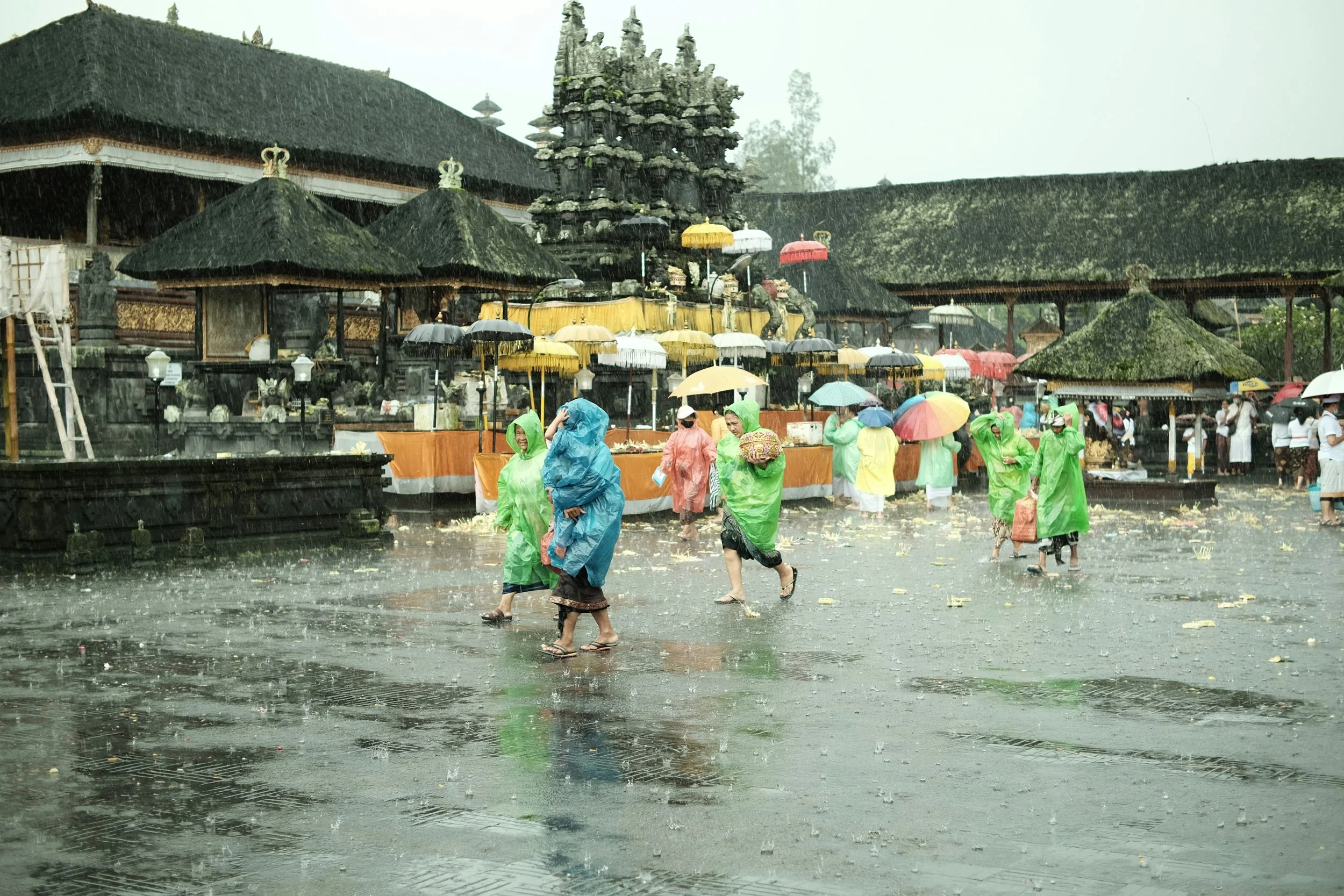 People wearing colorful rain ponchos and carrying umbrellas walk through a rain-soaked temple courtyard during a ceremony at a traditional Balinese temple in Bali, Indonesia.