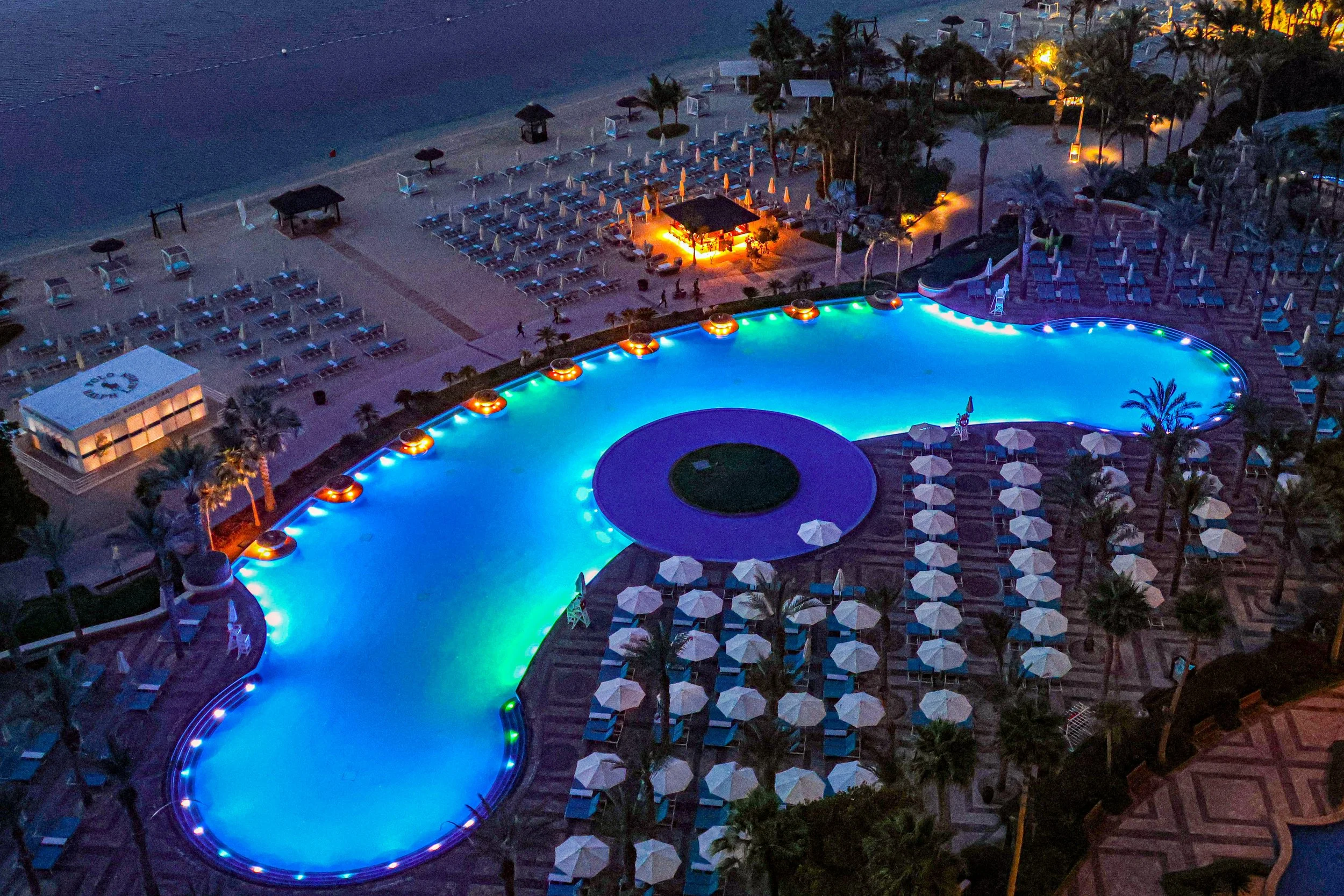 Aerial night view of a resort’s illuminated swimming pool with glowing blue and green lights, surrounded by sun loungers, umbrellas, and a beachfront area.