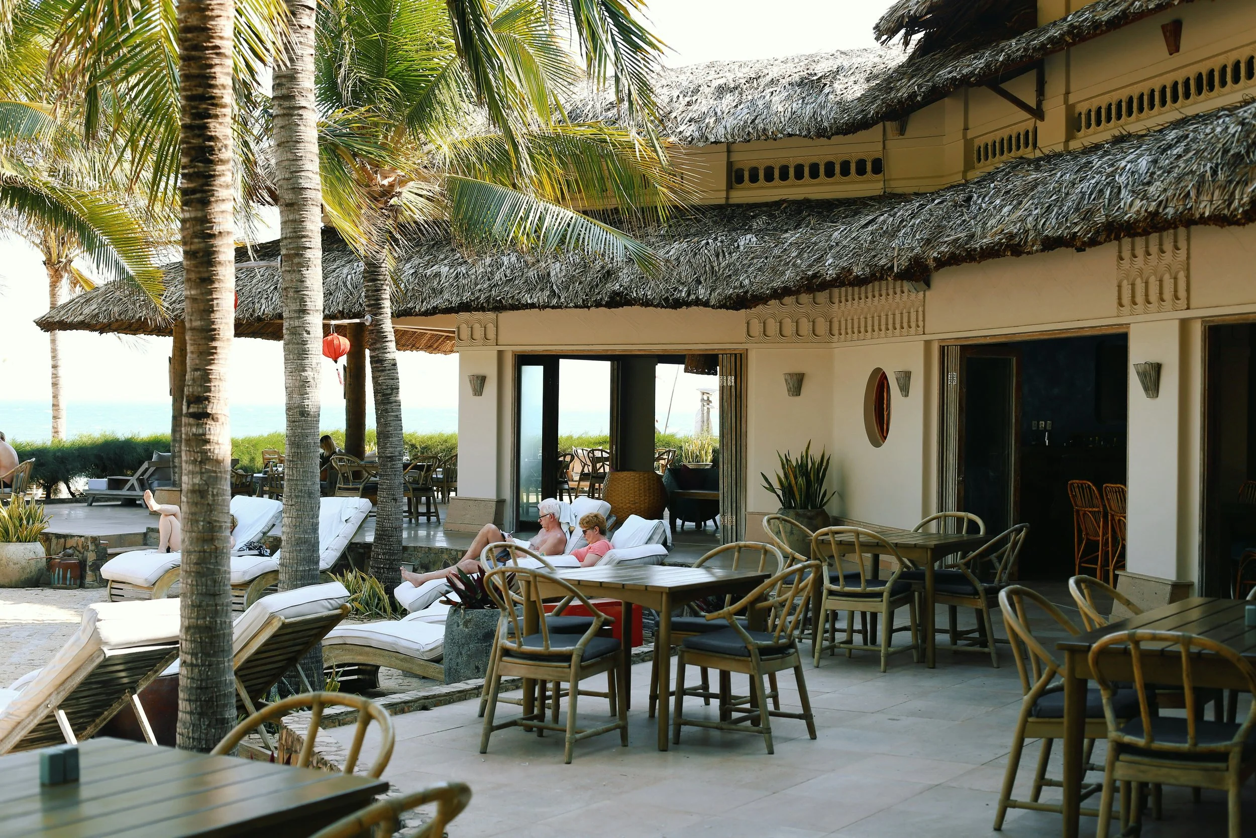 Outdoor lounge and dining area at a tropical beachfront resort, featuring palm trees, thatched roofs, sun loungers, and ocean views.