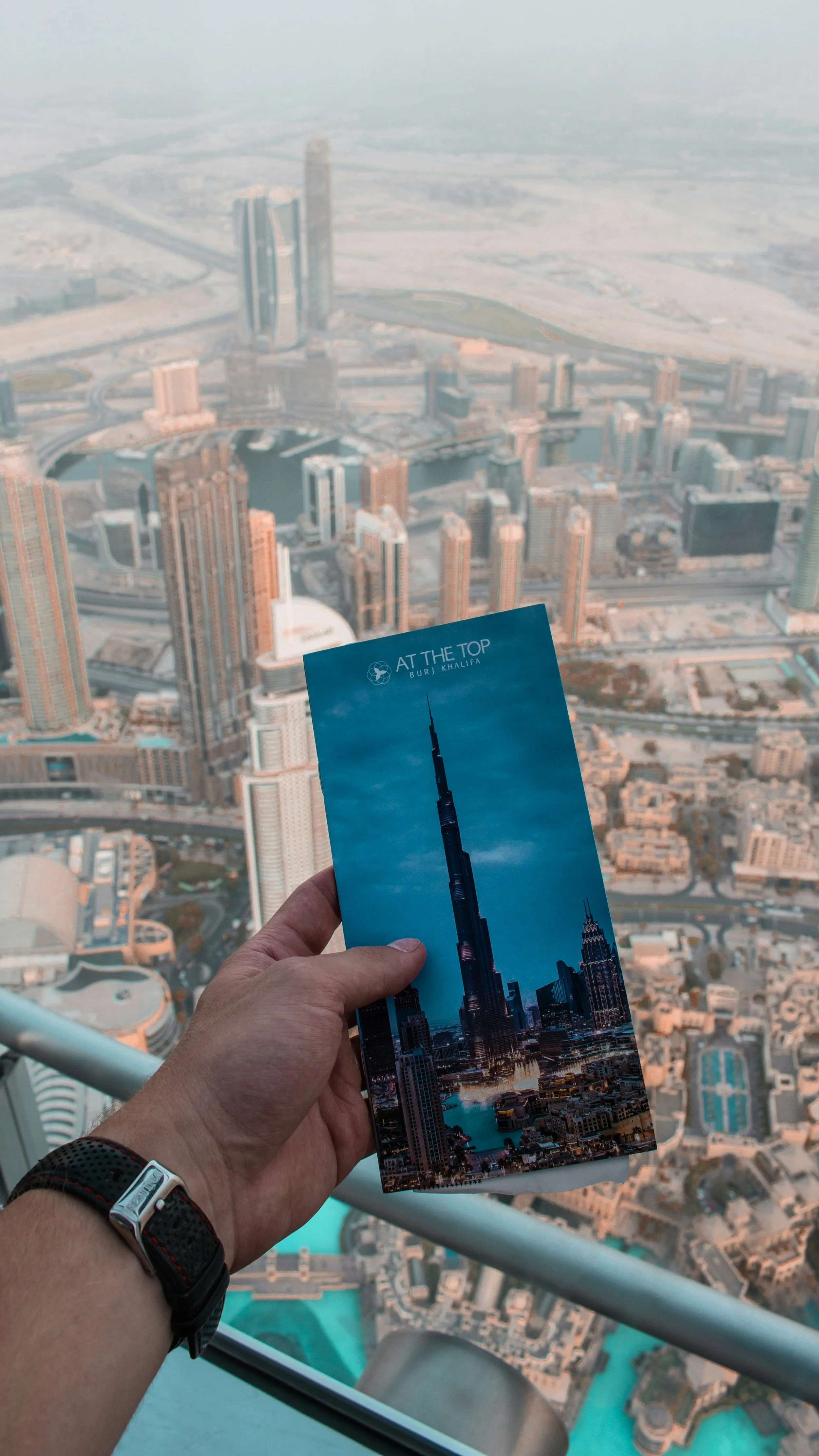 Hand holding an ‘At The Top Burj Khalifa’ booklet against the panoramic view of Downtown Dubai from the Burj Khalifa observation deck.