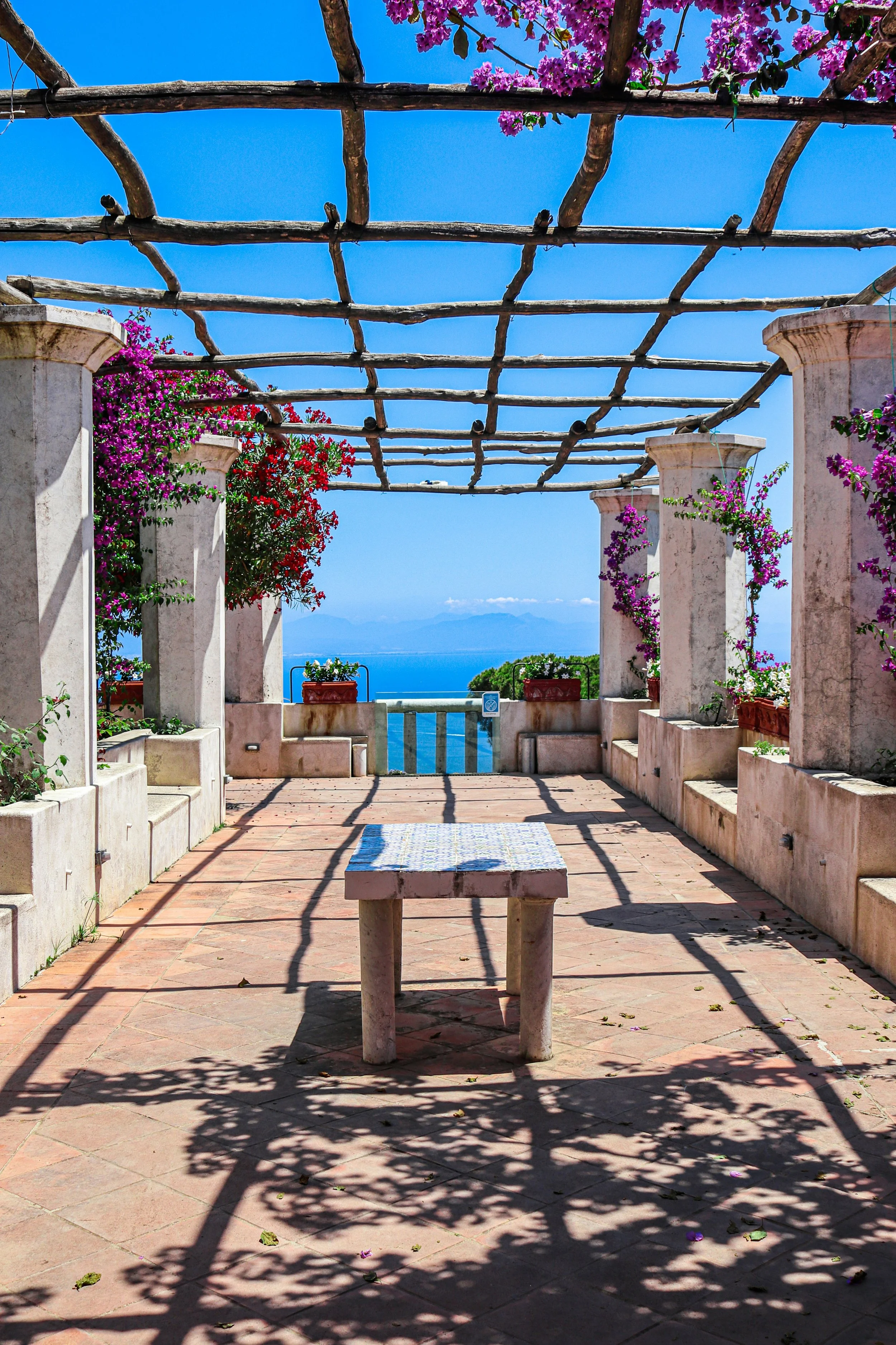 Sunlit pergola terrace on Italy’s Amalfi Coast with stone columns, flowering bougainvillea, and a clear view of the blue Mediterranean Sea.