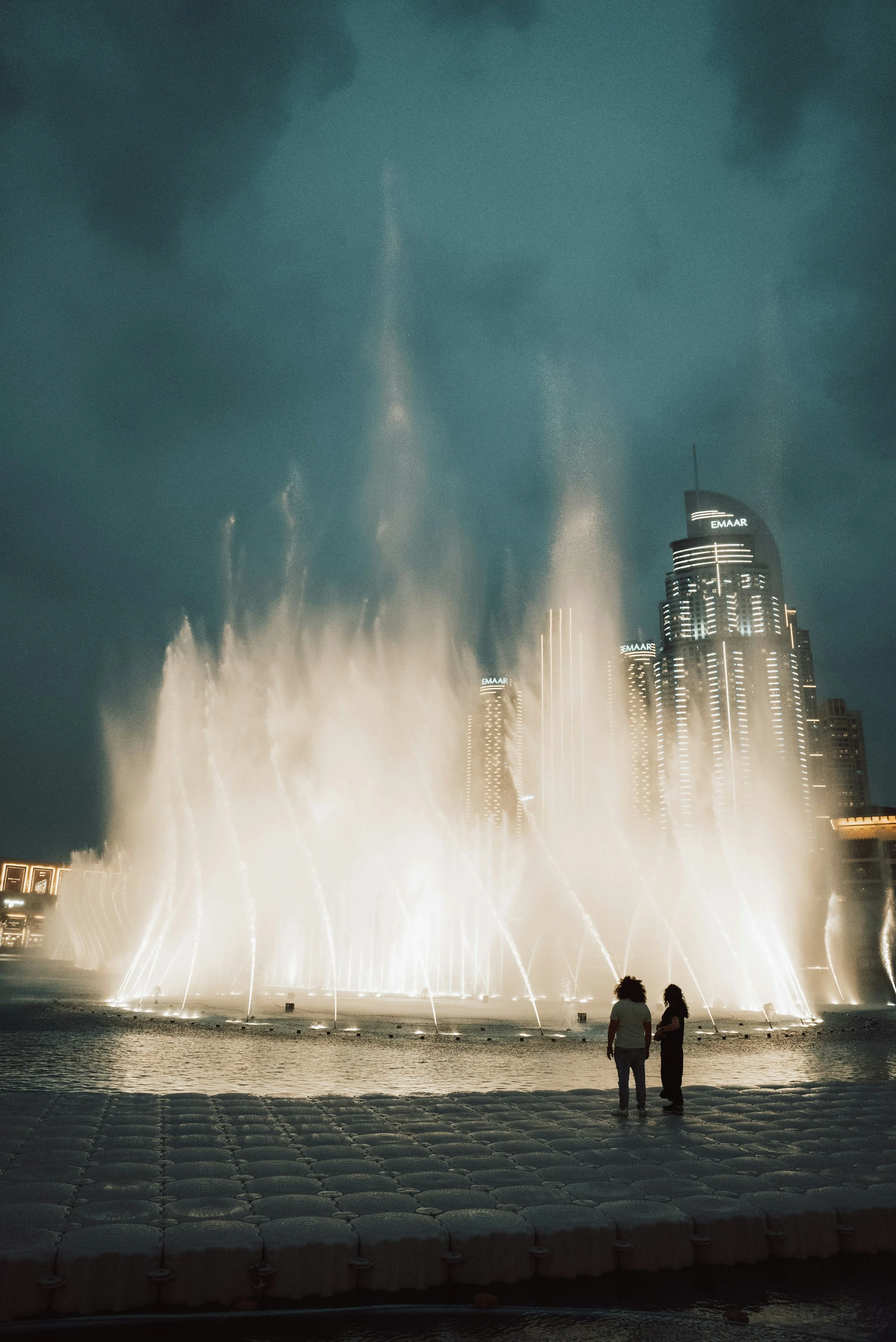 Two people watching the Dubai Fountain’s illuminated water show at night with the Address Downtown skyline in the background.