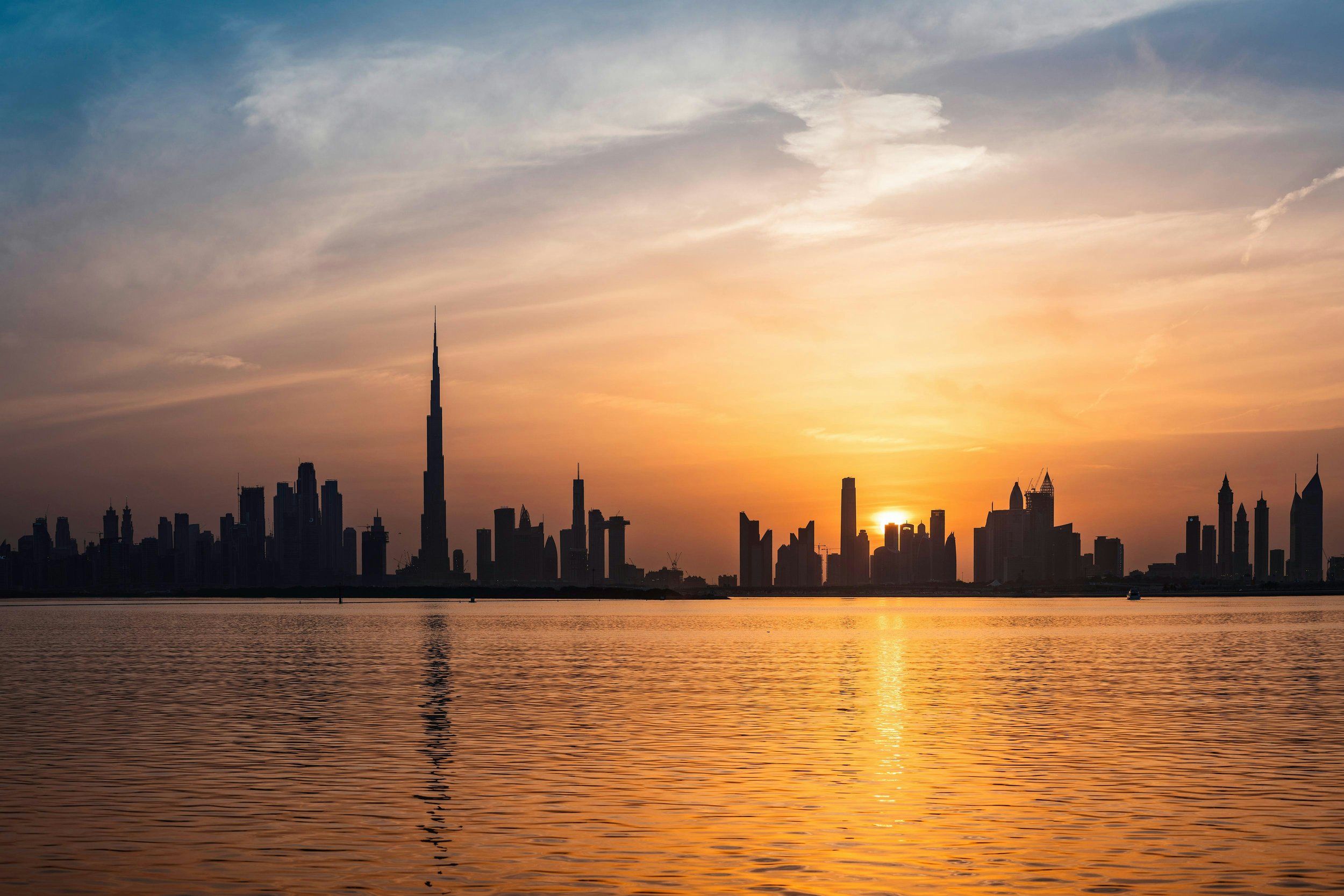 Dubai skyline at sunset with the Burj Khalifa silhouetted against an orange sky, reflected over calm water.