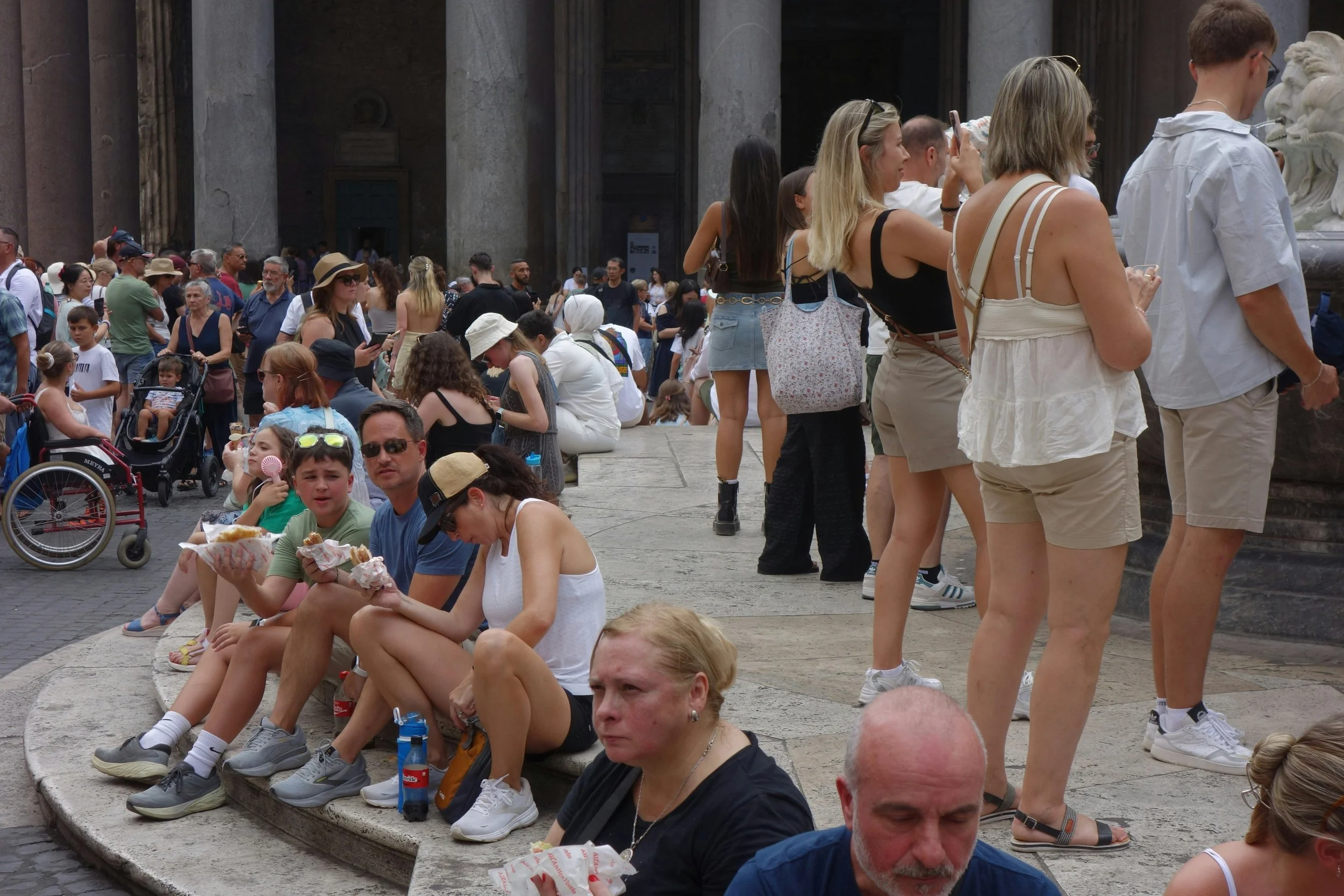 A crowded tourist area with groups of people sitting on stone steps, eating snacks, taking photos, and gathering near a historic landmark.