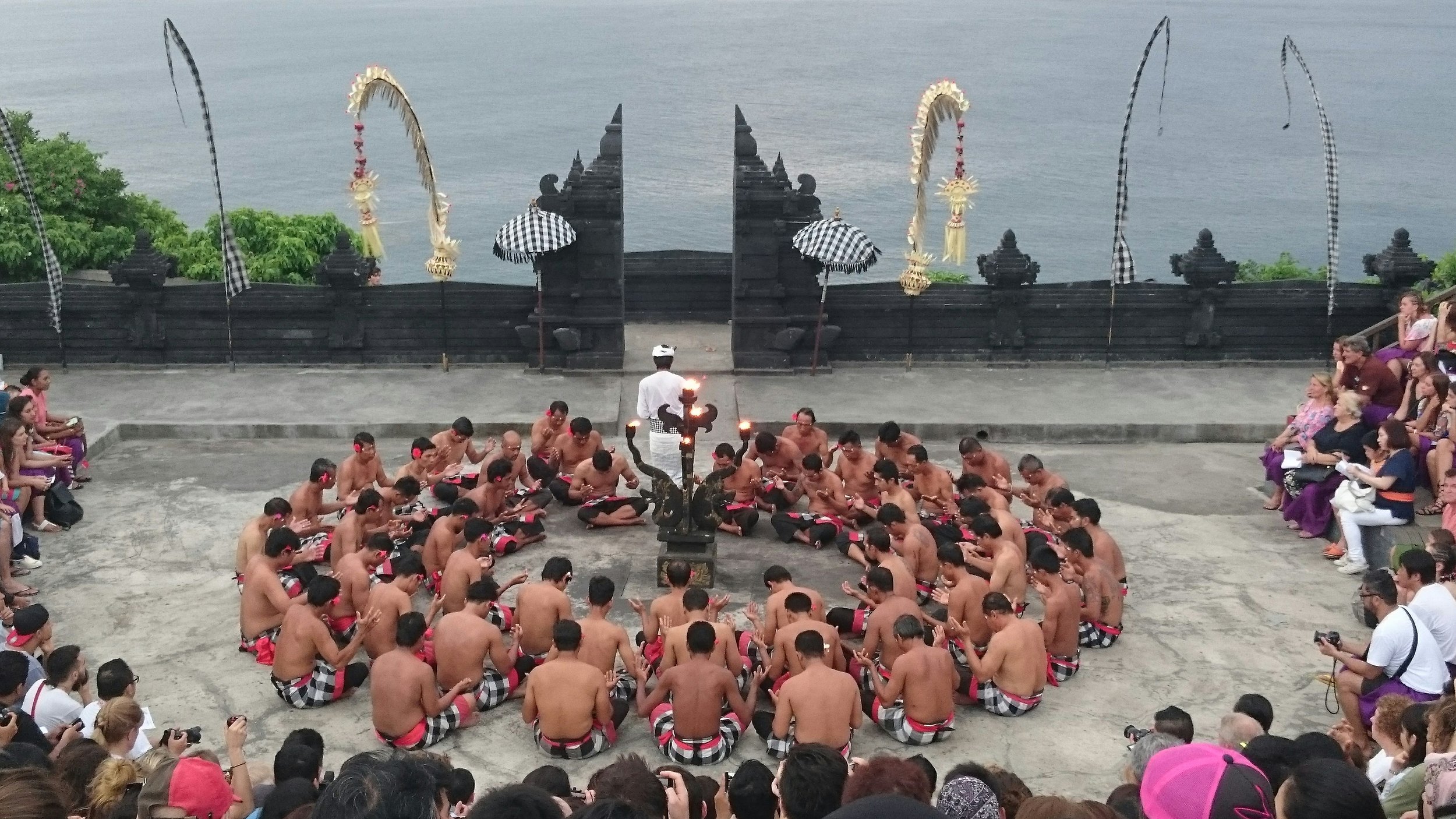 Traditional Kecak fire dance performance in Bali with dozens of performers seated in a circle at an open-air temple overlooking the ocean, watched by an audience.