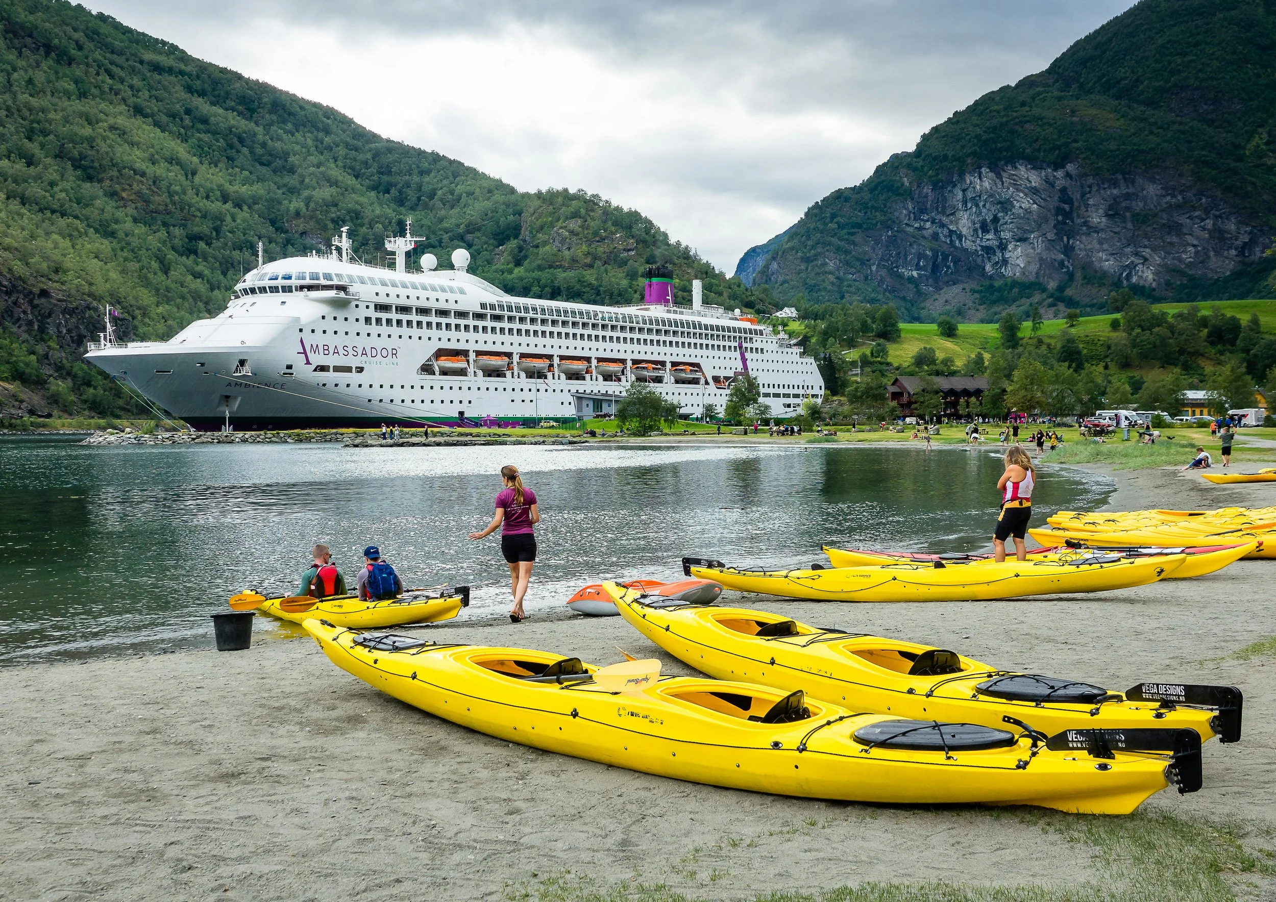 Cruise ship docked in a scenic Norwegian fjord with yellow kayaks on the shoreline and mountains in the background.