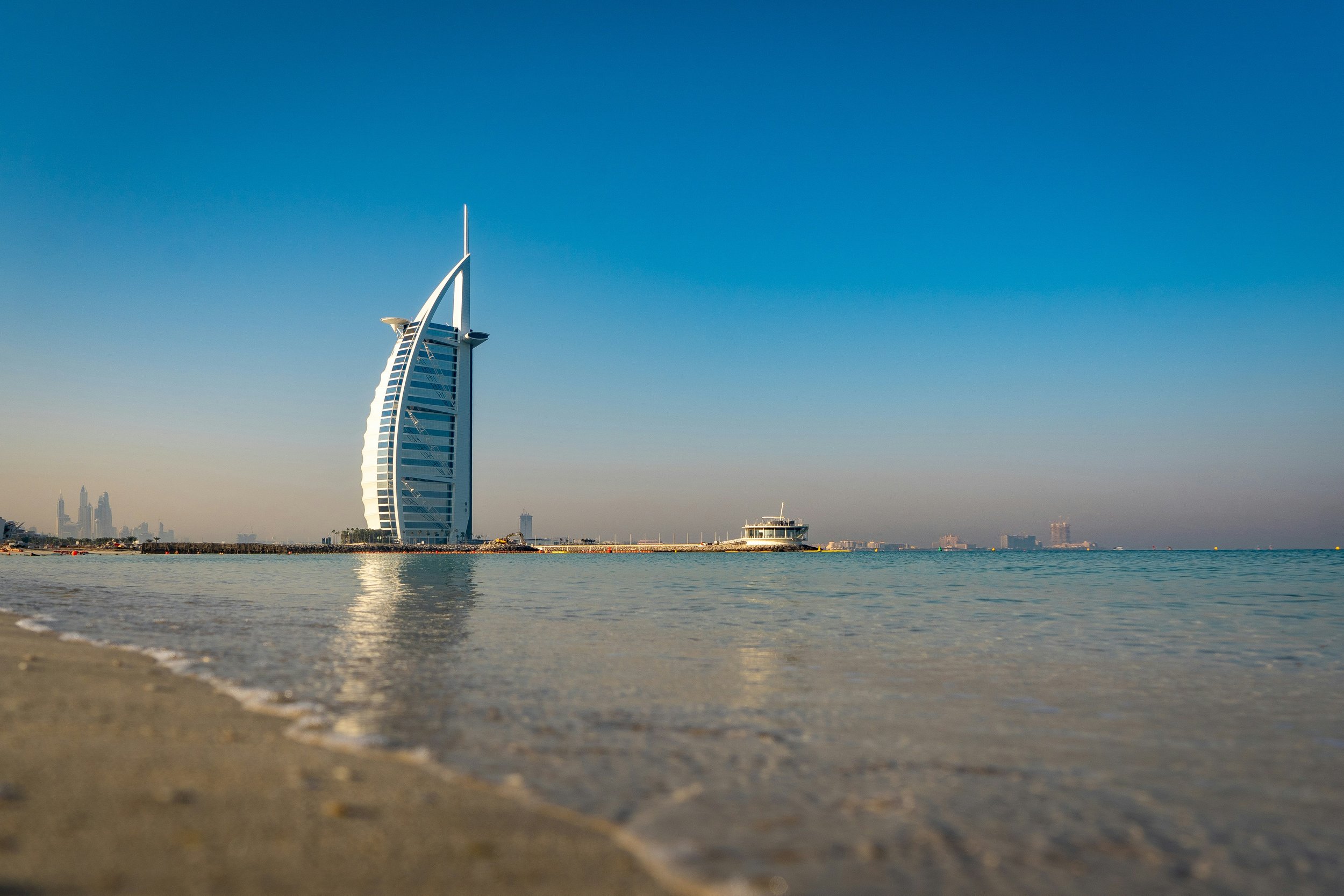 View of the Burj Al Arab from the shoreline in Dubai, with calm blue water and a clear sky.