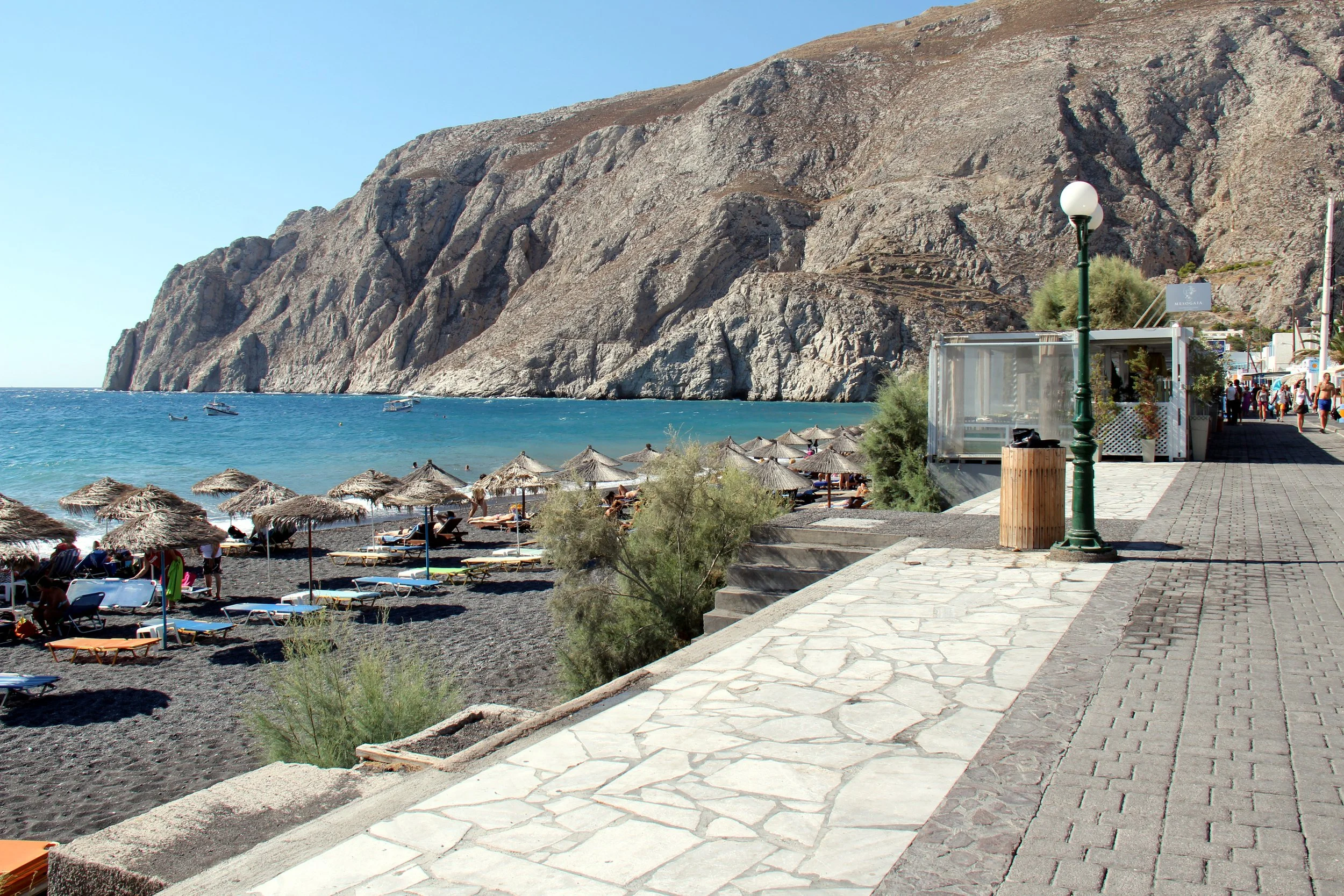Kamari Beach in Santorini with black sand, sun loungers, thatched umbrellas, clear blue water, and a seaside promenade along the cliffs.