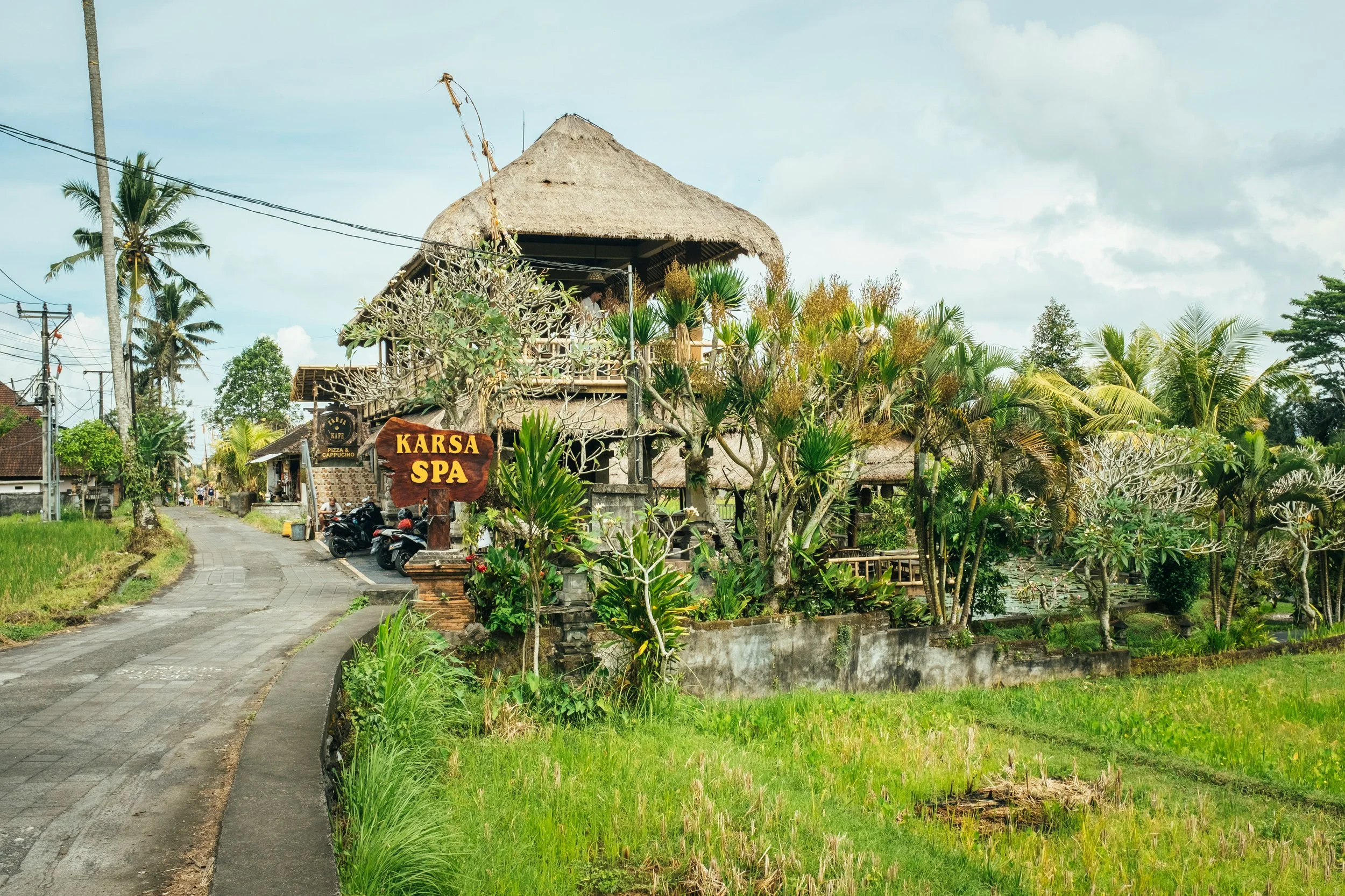 Traditional Balinese spa building with a thatched roof along a quiet roadside in Ubud, surrounded by lush greenery, palm trees, and rice fields.