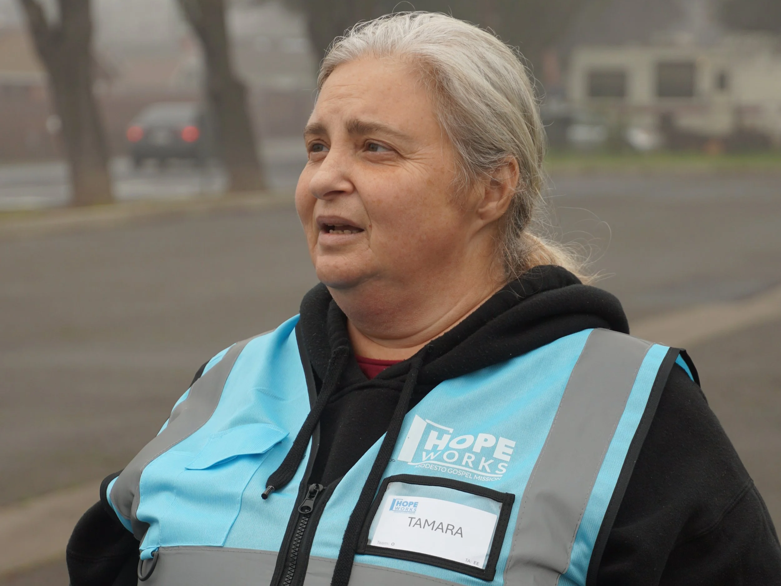 A woman with gray hair wearing a black hoodie and a light blue vest with the logo 'Hope Works' and a name tag that says 'Tamara.' She appears to be outdoors on a cloudy day.