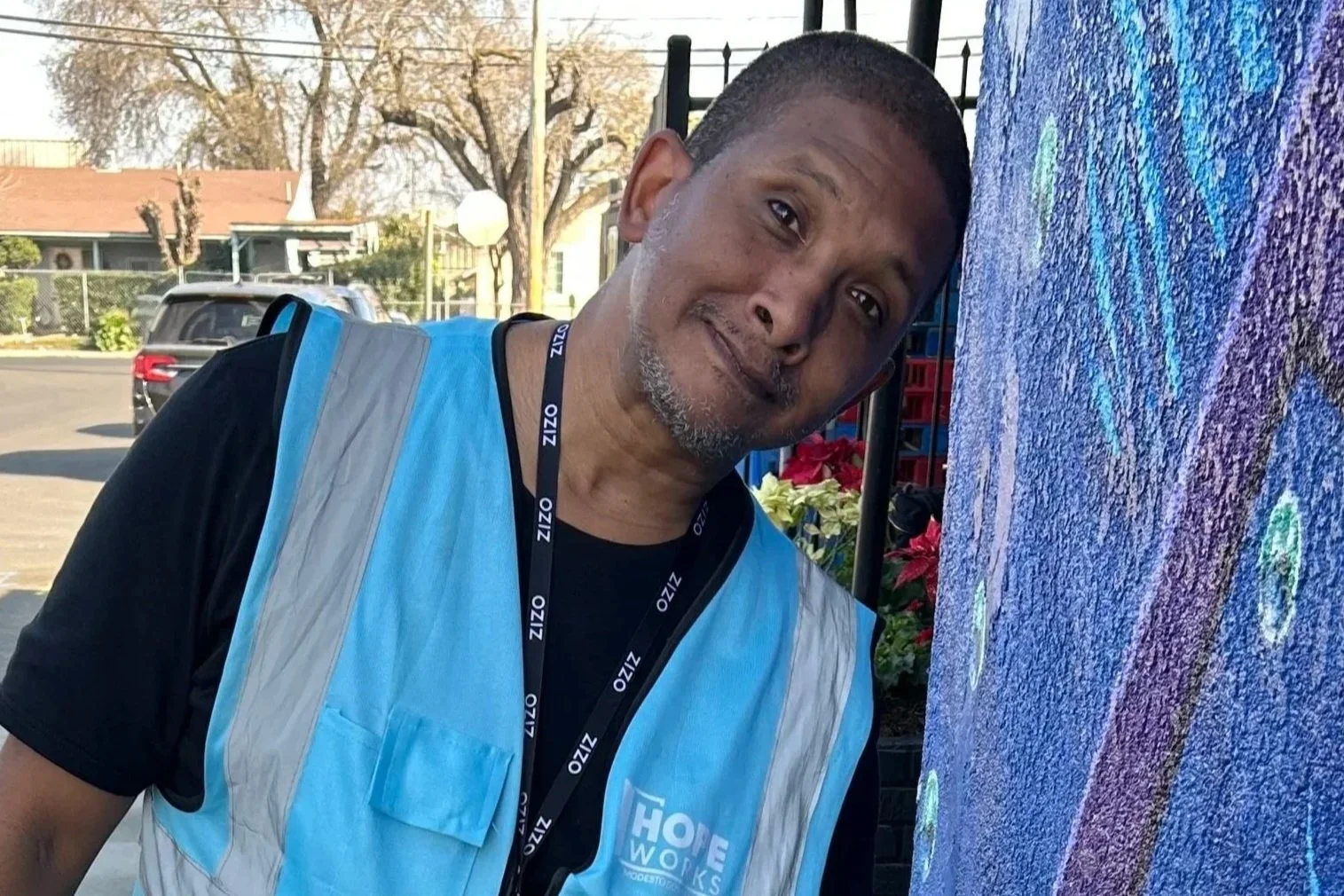 A man with a gray beard and short hair wearing a black shirt and a light blue reflective vest, standing next to a colorful mural on a wall, outdoors during daytime.