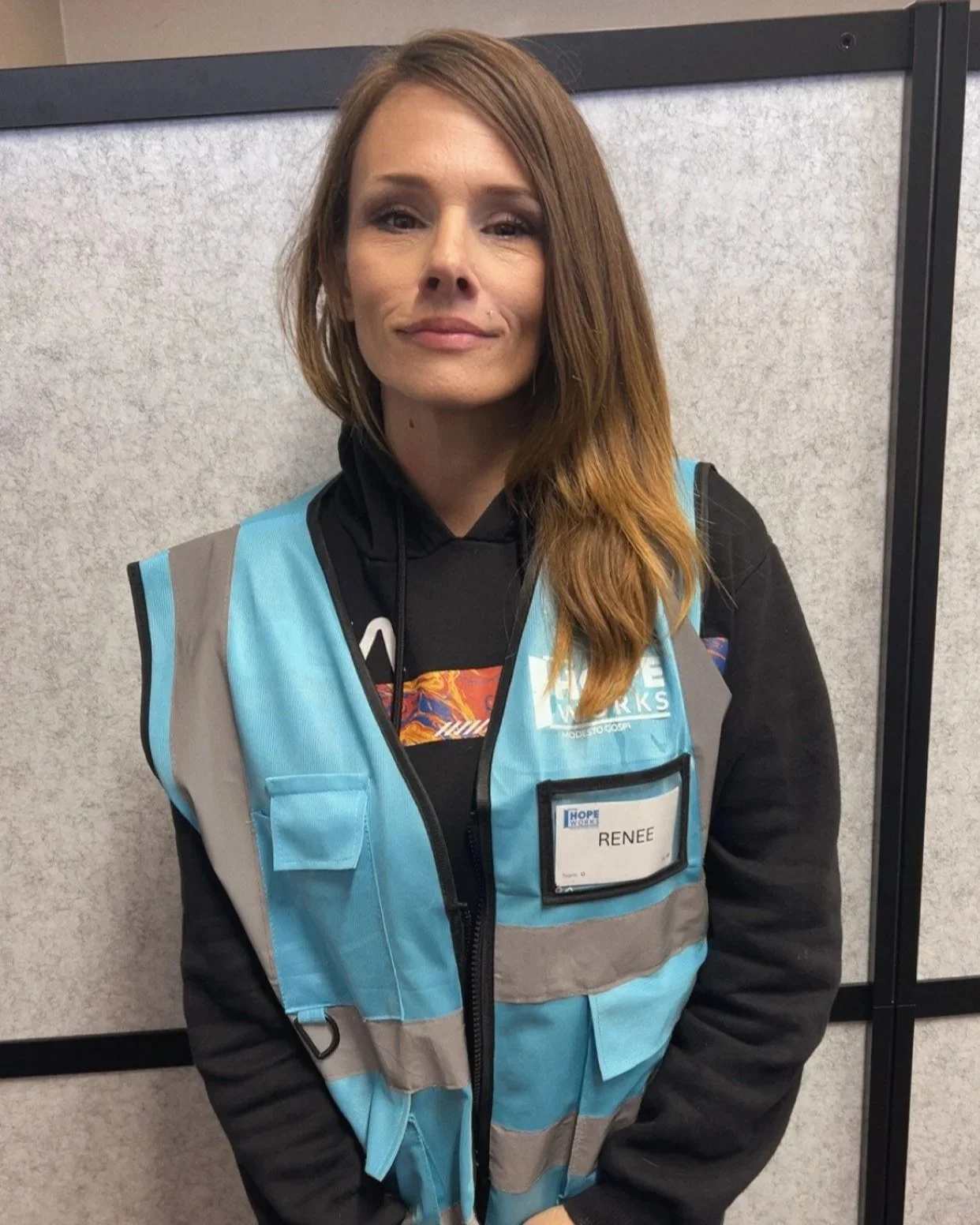 A woman with red hair wearing a blue safety vest and black hoodie, standing indoors against a gray partition. She has a name tag that reads 'Renee'.