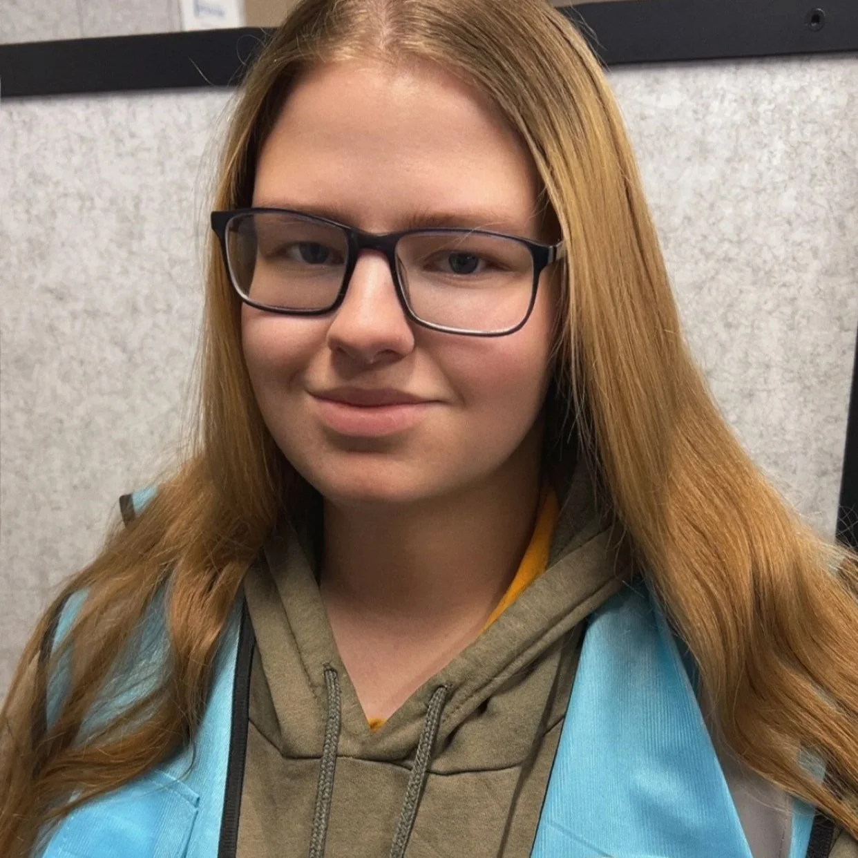 A young woman with long red hair, glasses, and a light brown hoodie, smiling slightly, standing indoors against a neutral background.