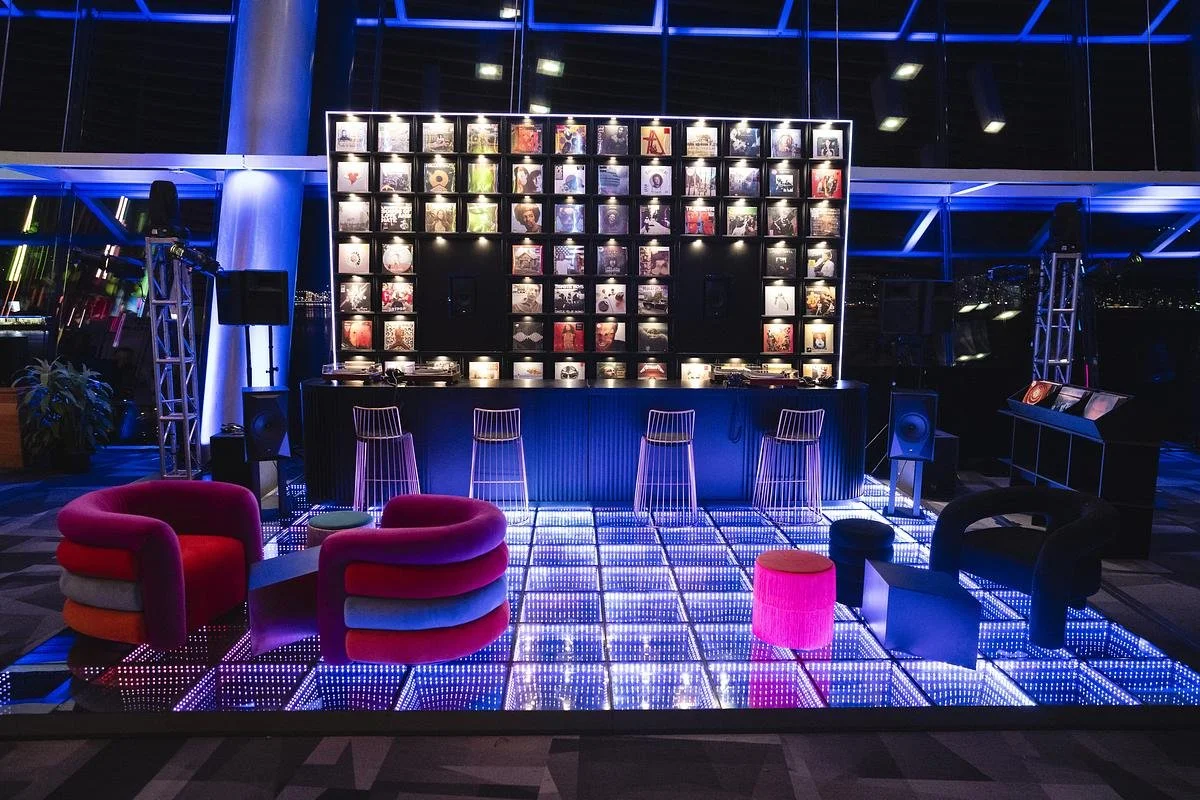 Nightclub or lounge with a illuminated dance floor, colorful chairs, a bar with a backdrop of framed photographs, and city lights visible through large windows.