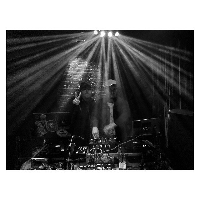 Two DJs in a dark club with stage lights and a brick wall behind them, operating DJ equipment.