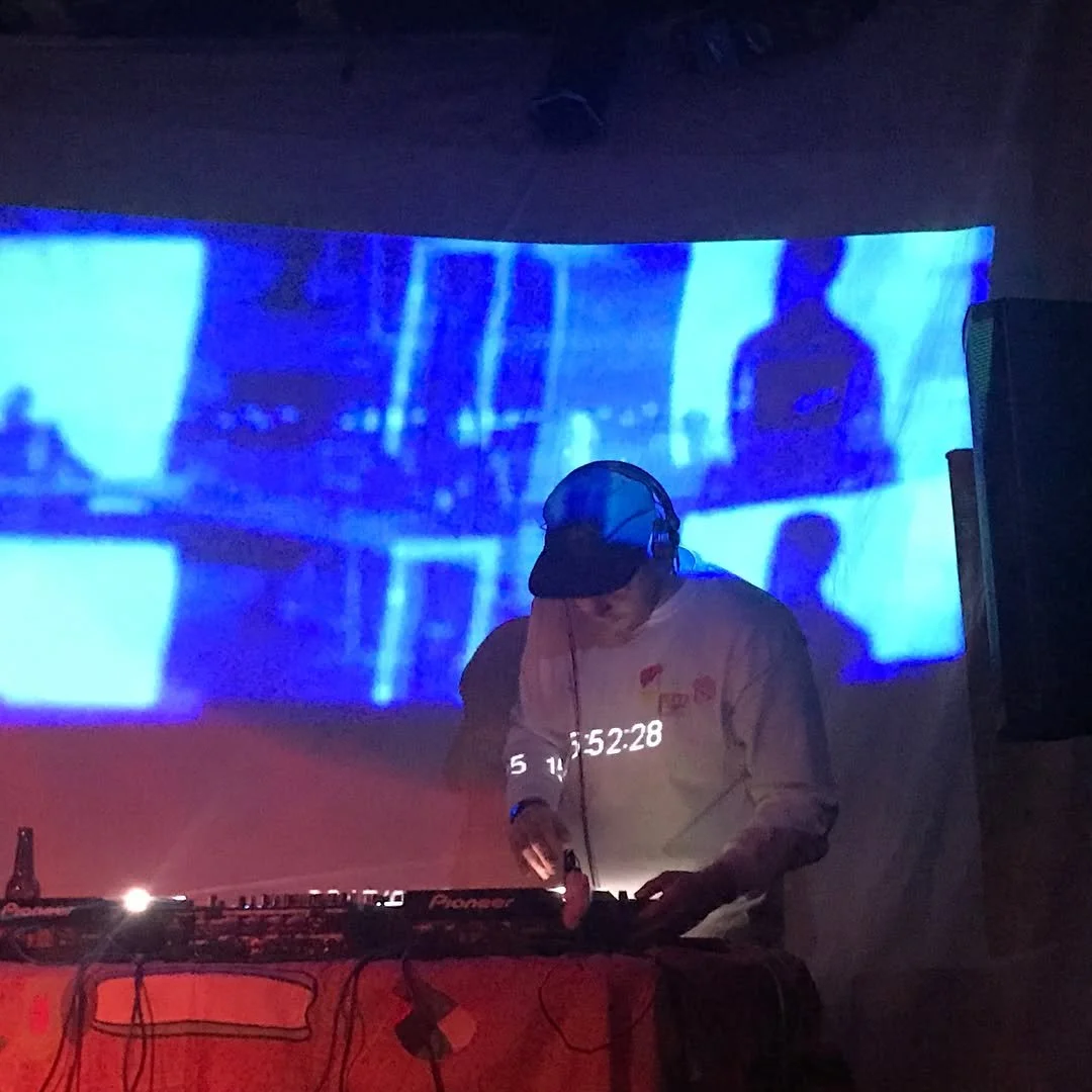 DJ wearing headphones and a cap, DJing at a nightclub with a blue abstract video projected on the wall behind him.