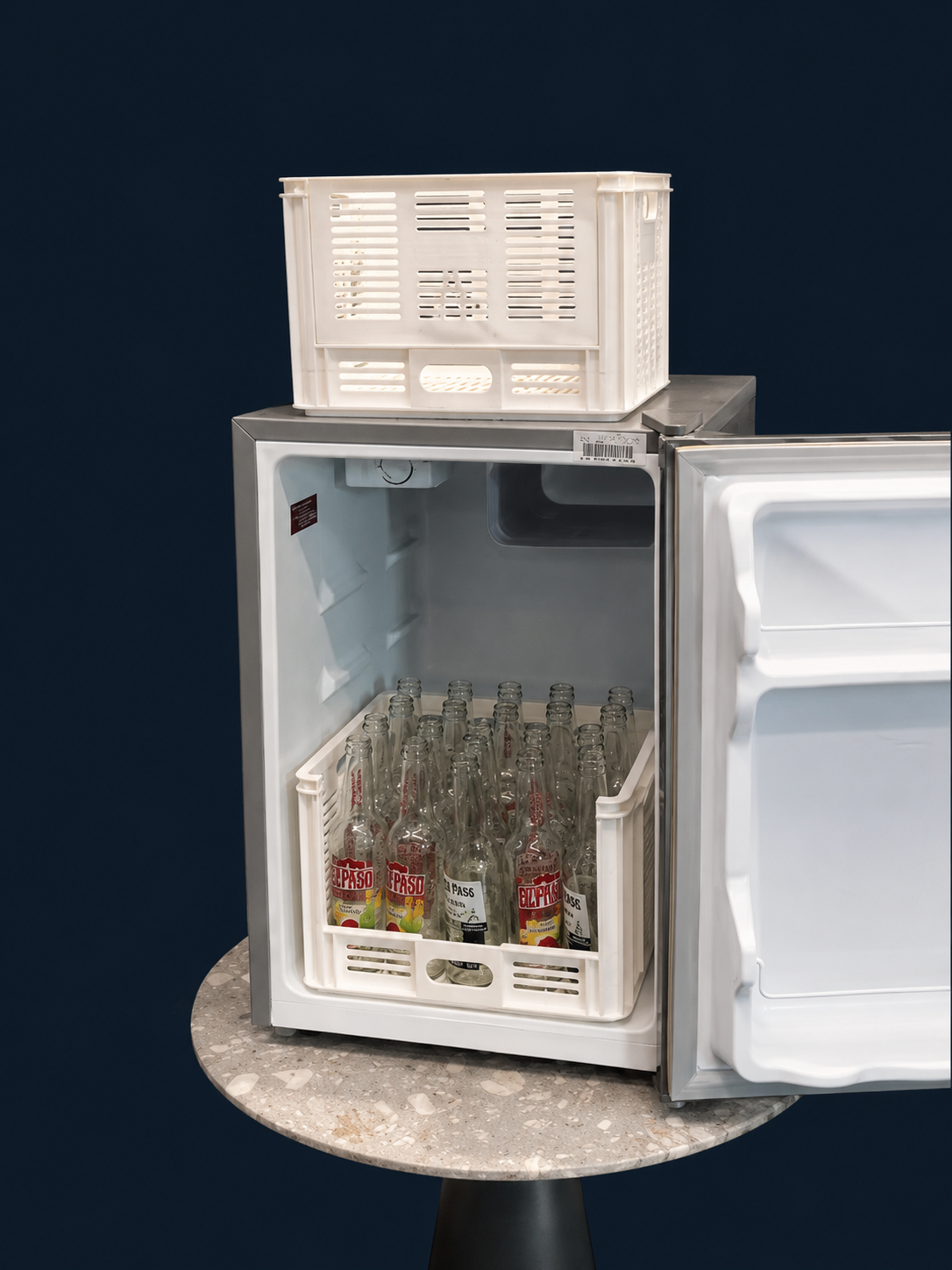 Small refrigerator with an open door, containing glass bottles of Paso soda and a plastic crate on top, set on a round table against a dark background.