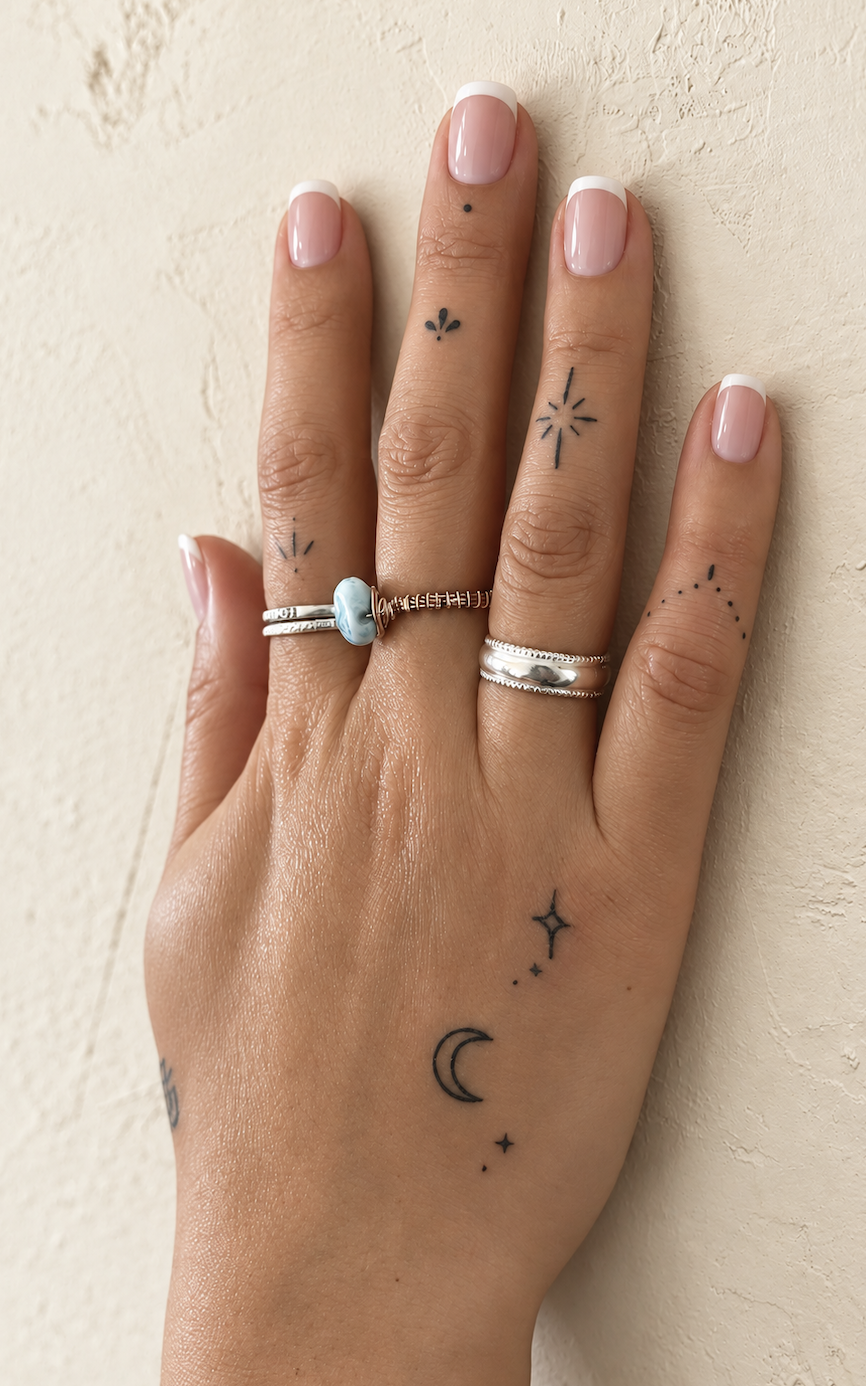 A hand with light pink manicured nails and multiple silver rings, resting against a textured beige wall, featuring various small tattoos including moons, stars, and geometric designs.