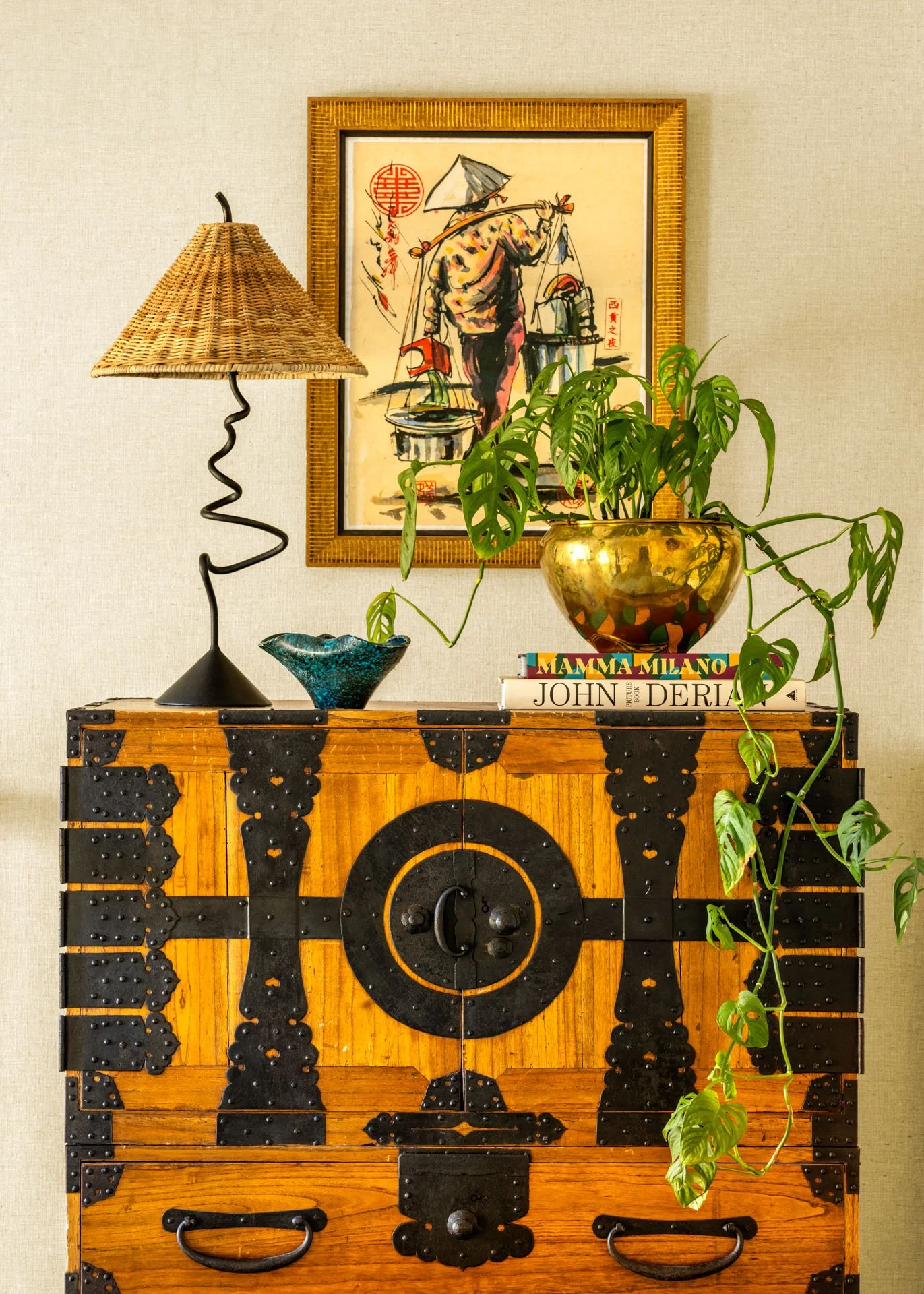 A wooden cabinet with black decorative metal accents, a table lamp with a woven shade, a green potted plant in a gold pot, a small decorative bowl, and books titled "Mamma Milano" and "John Derian" are arranged on top. A framed Asian-style painting of a person carrying a load, with traditional Chinese red seals, hangs on the beige wall behind the cabinet.