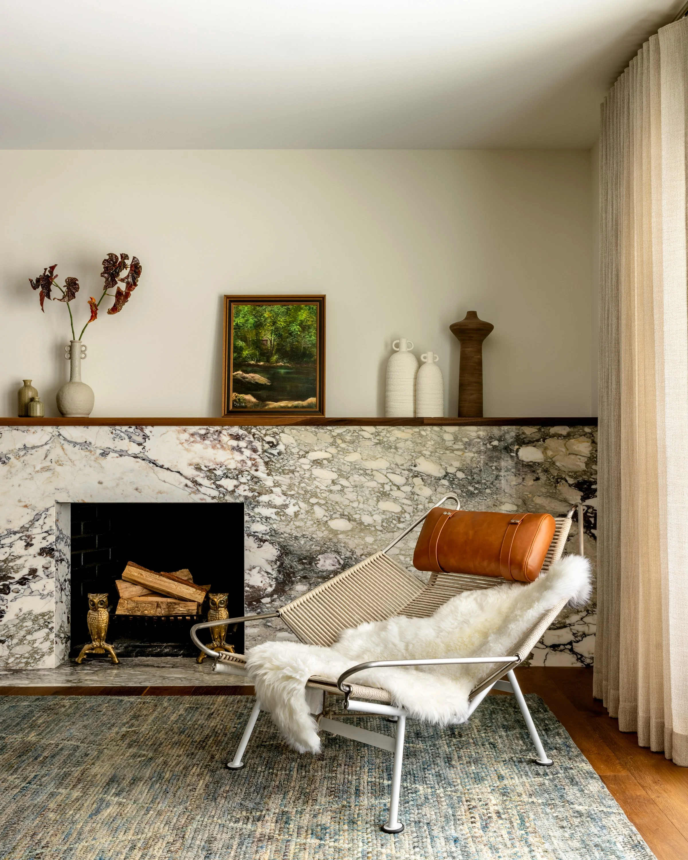 Living room with a marble fireplace, cozy chair with sheepskin throw, and decorative vases and artwork on the mantel