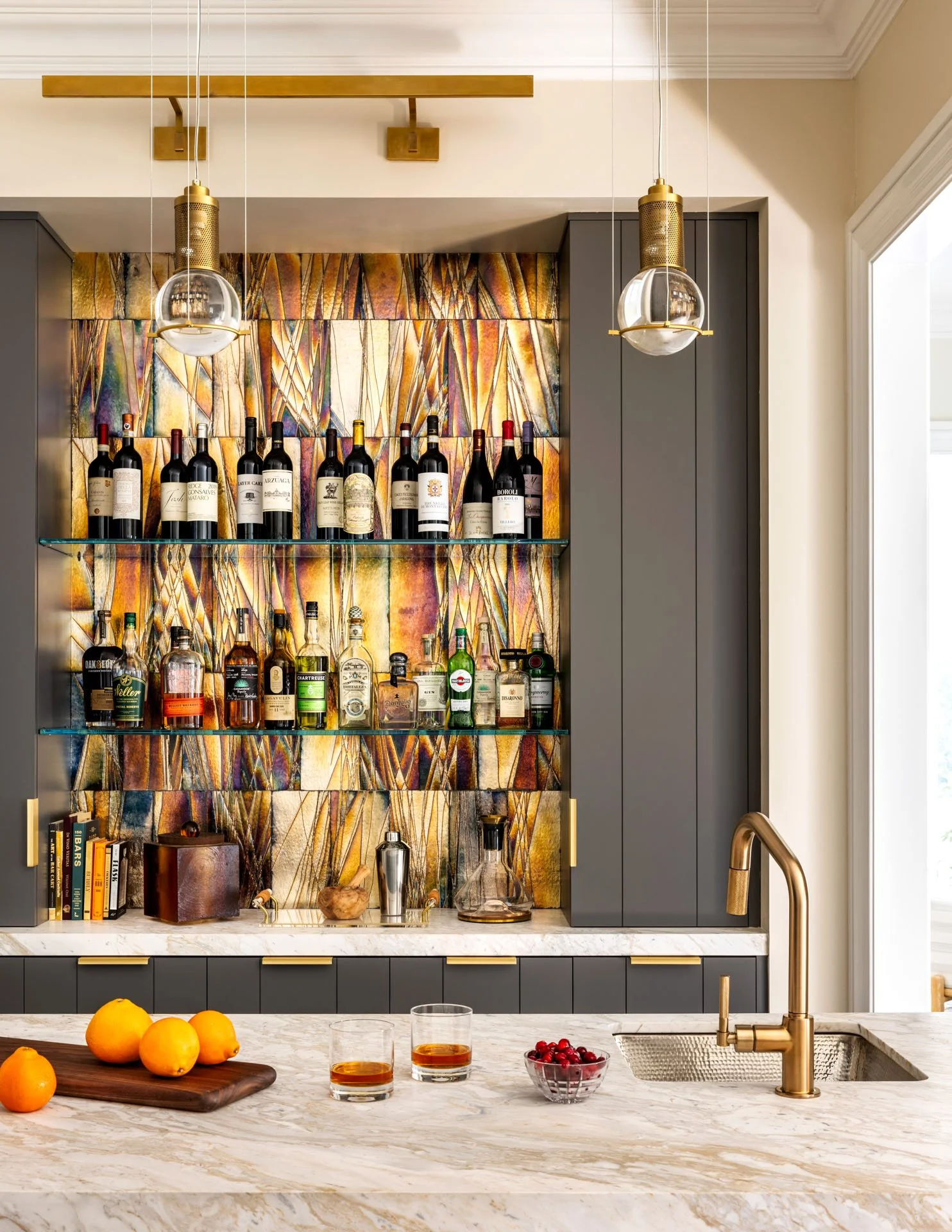Kitchen bar area with a marble countertop, glasses of whiskey, a bowl of cranberries, oranges, a sink with a gold faucet, shelves with various bottles of alcohol, decorative books, and a colorful abstract backsplash.